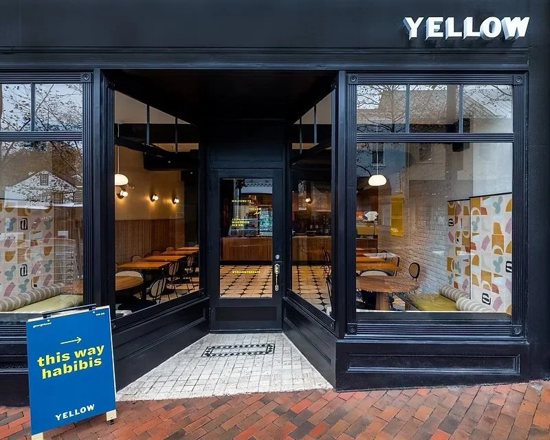 Exterior of a restaurant or cafe with large glass windows and a glass door. The sign on top says "YELLOW". There is a blue sandwich board outside with yellow and white text that reads "this way habibis" with a yellow arrow pointing in a direction.