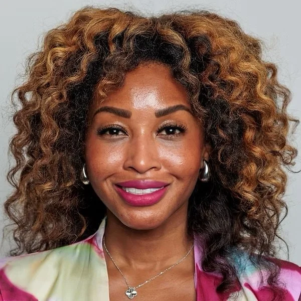 Close-up portrait of a woman with curly, voluminous hair, wearing pink lipstick, hoop earrings, and a colorful top, smiling at the camera.