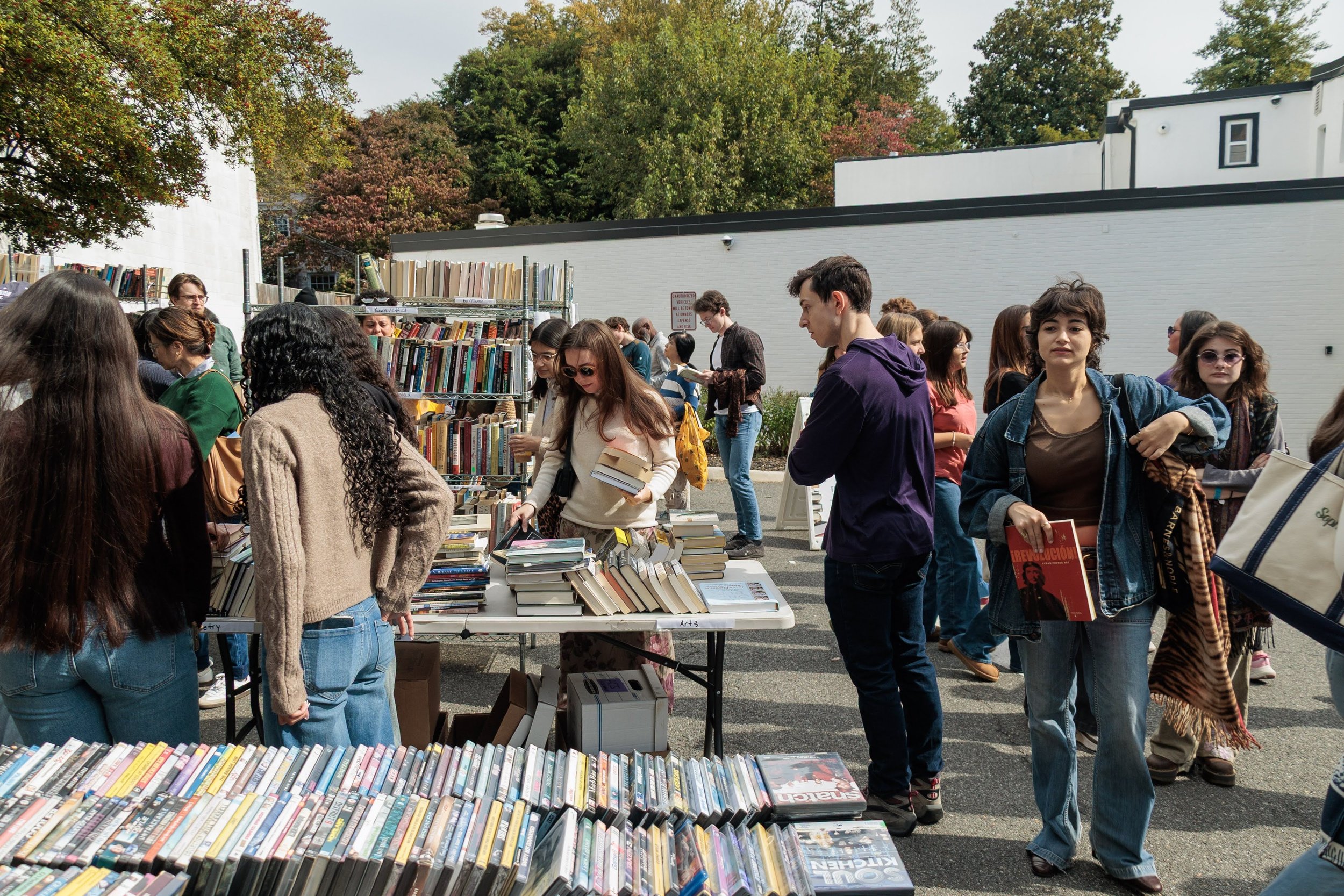 The photo shows a parking lot converted into a pop up market. There are many people looking at books on tables and racks, some appear to have made selections. The atmosphere is joyful.