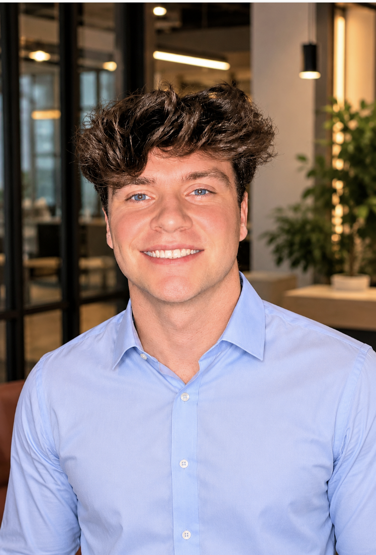 Portrait of a young man with curly brown hair and blue eyes, smiling, wearing a light blue button-up shirt, in a modern indoor setting.