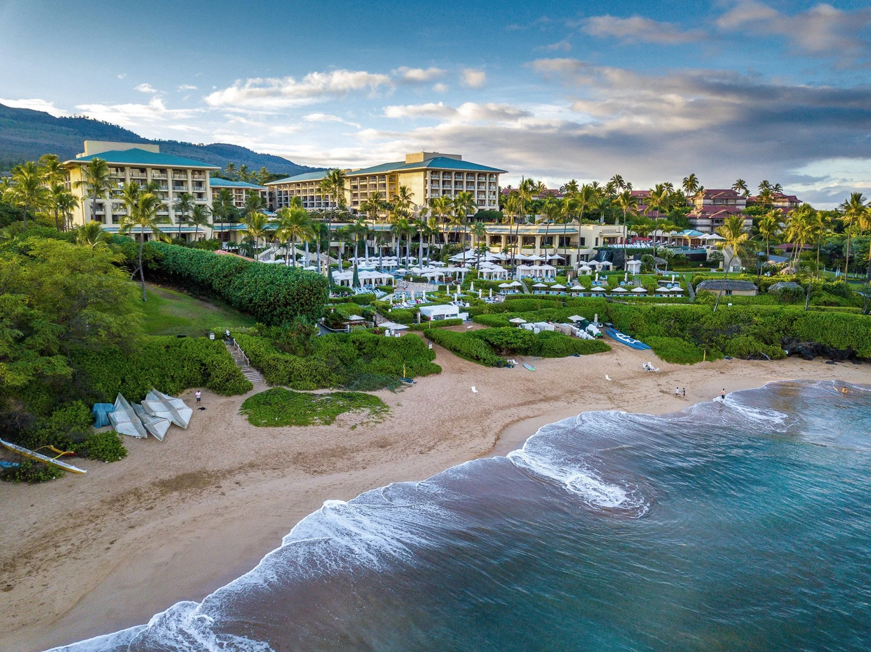 A large beachfront resort with multi-story buildings, lush greenery, numerous white umbrellas and lounge chairs on a patio, a sandy beach, and ocean waves. Four Seasons Maui Resort