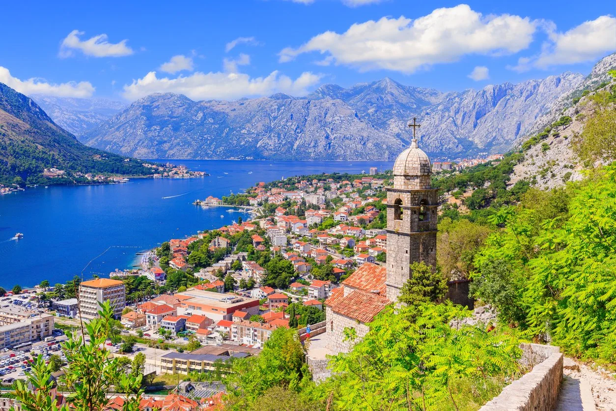 A scenic view of a town with red-roofed buildings along a large lake, surrounded by green hills and mountains under a partly cloudy blue sky. Montenegro