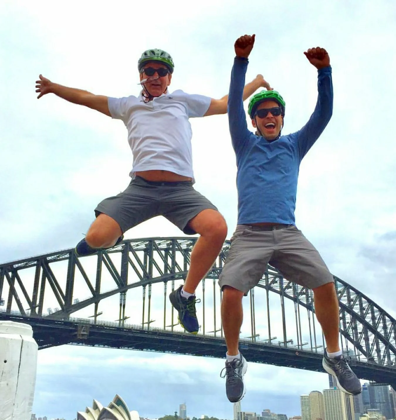 Two men are jumping in the air in front of the Sydney Harbour Bridge and the Sydney Opera House, both wearing helmets and sunglasses, with a city skyline in the background.