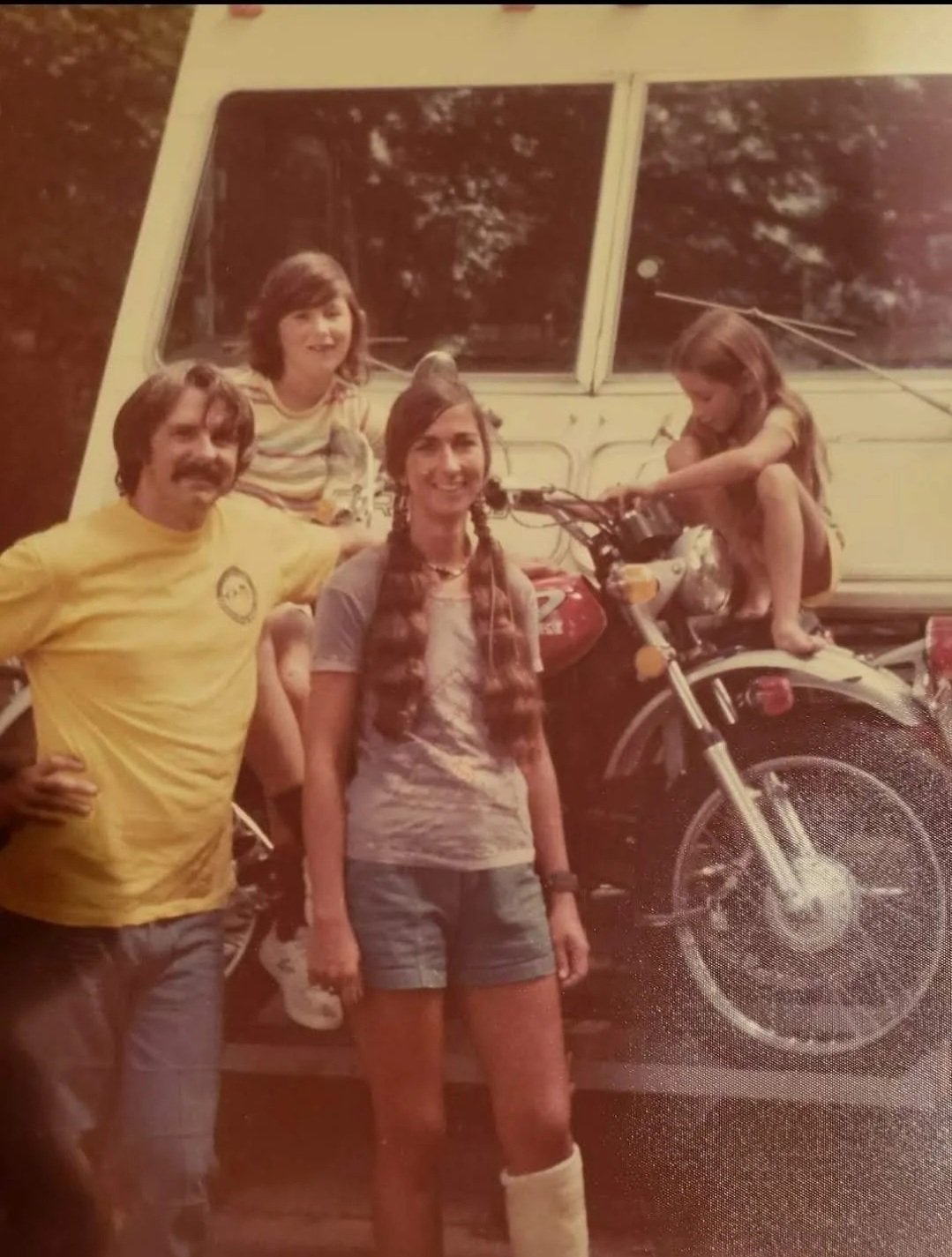 A vintage photograph of a family outdoors with a motorcycle. Two are sitting on the motorcycle, one on the seat and the other on the handlebar, while a man and a woman stand beside them. The man is wearing a yellow shirt and the woman has long braided hair and is wearing a casual outfit. The background features trees and a large garage door.