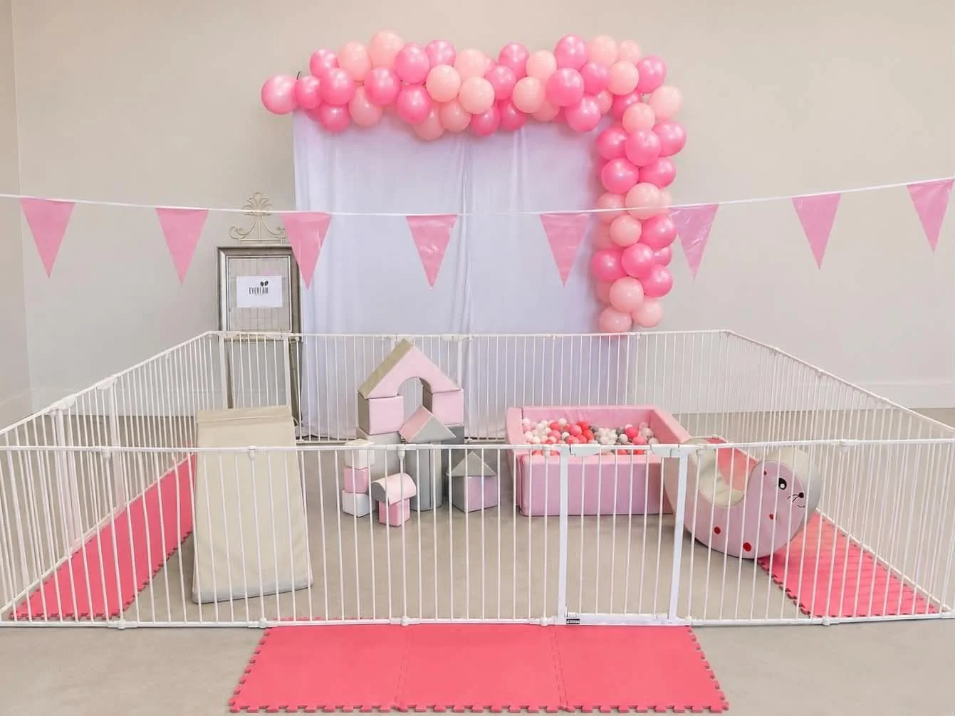 Soft pink and peach balloons arranged in an arch and cascade against a white backdrop, with pink triangular bunting banners hanging across a beige wall. A playpen with pink and gray padded mats, soft blocks, and a ball pit is set up inside, suitable for a child's party or play area.