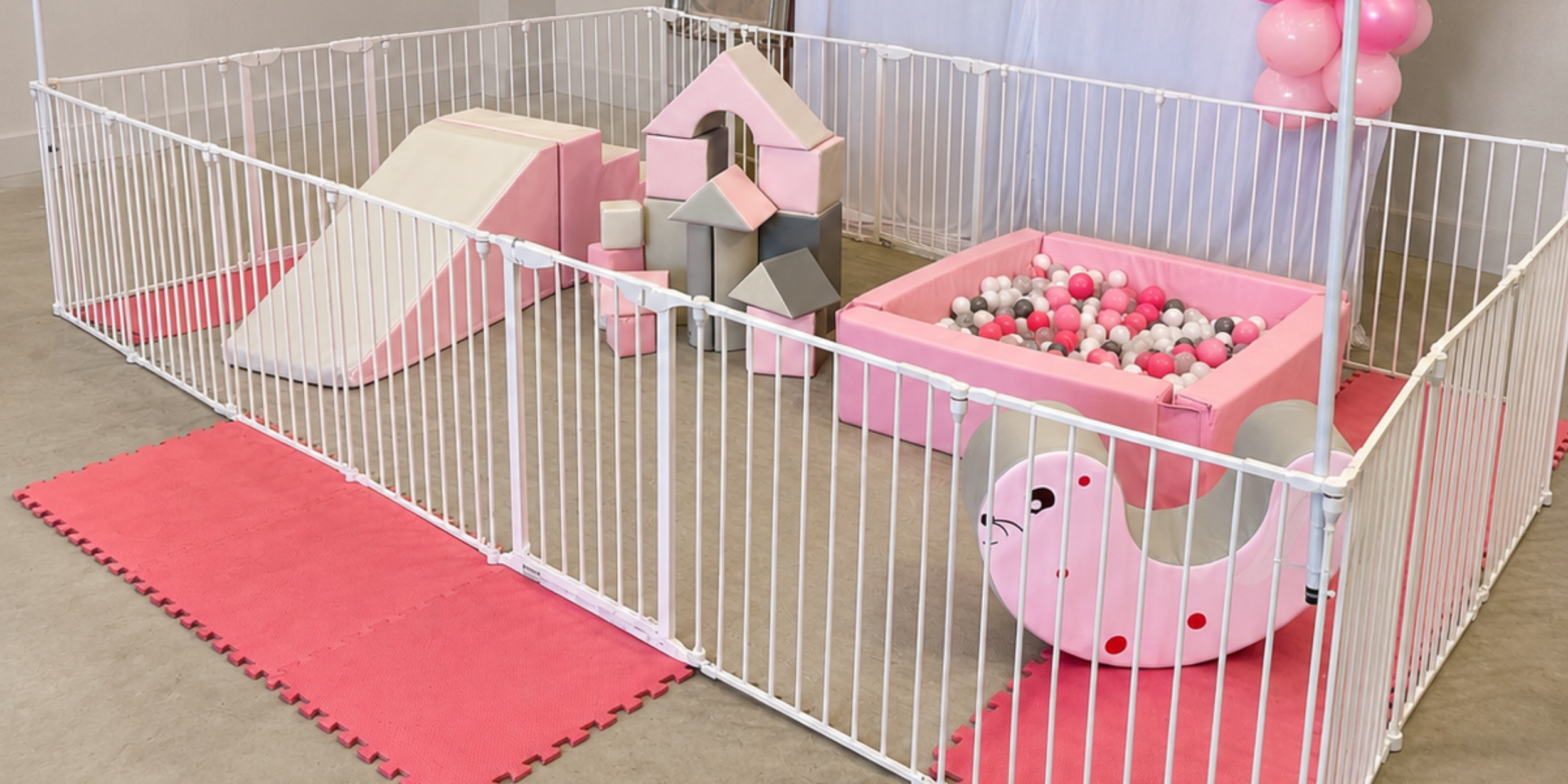 A small indoor playpen with a pink and white theme, featuring a slide, foam climbing blocks, and a ball pit filled with pink, white, and gray balls. Pink balloons are tied to a stand in the background.