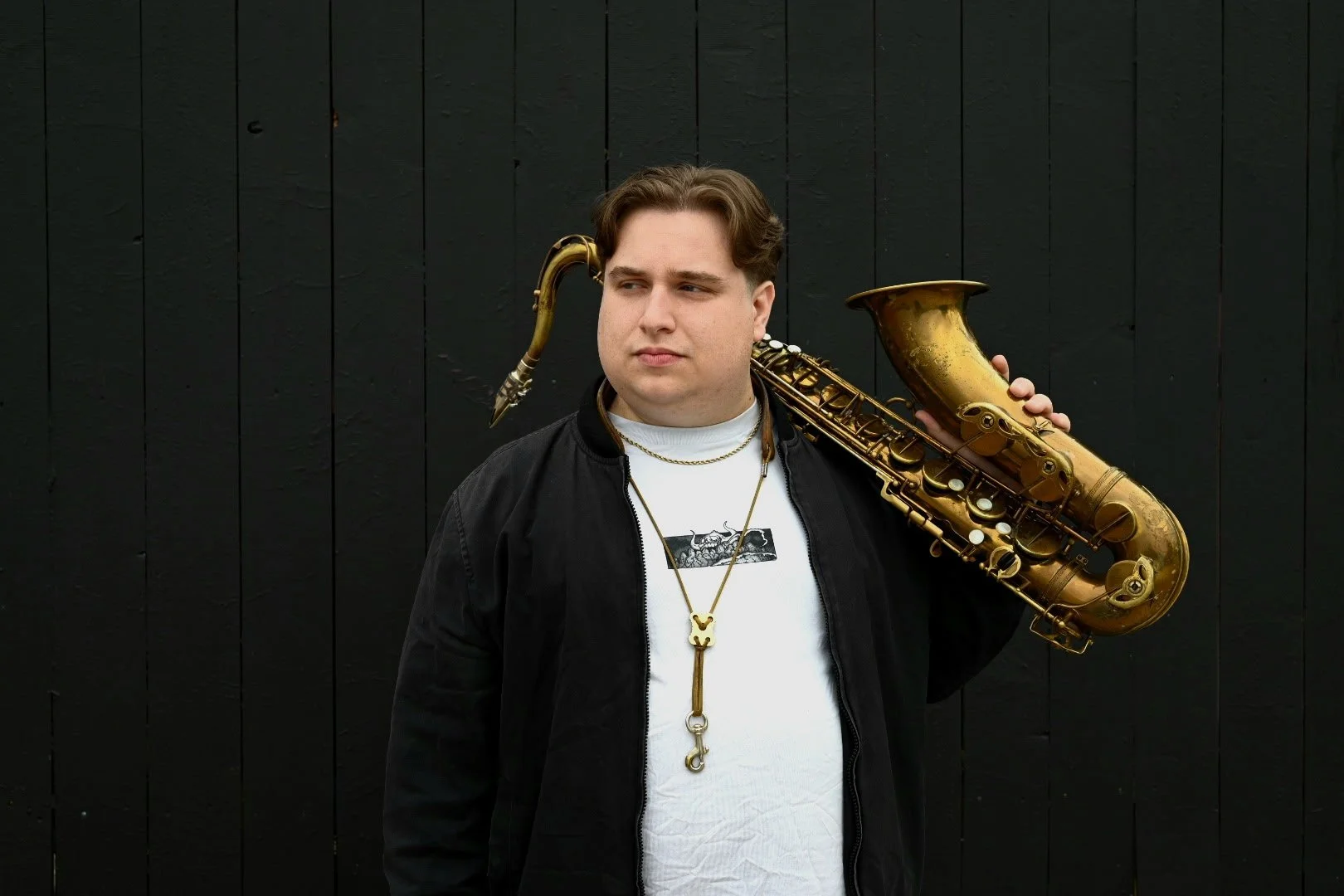 Young man holding a gold saxophone on shoulder, wearing a black jacket, necklace, and neck strap, standing against black wooden background.