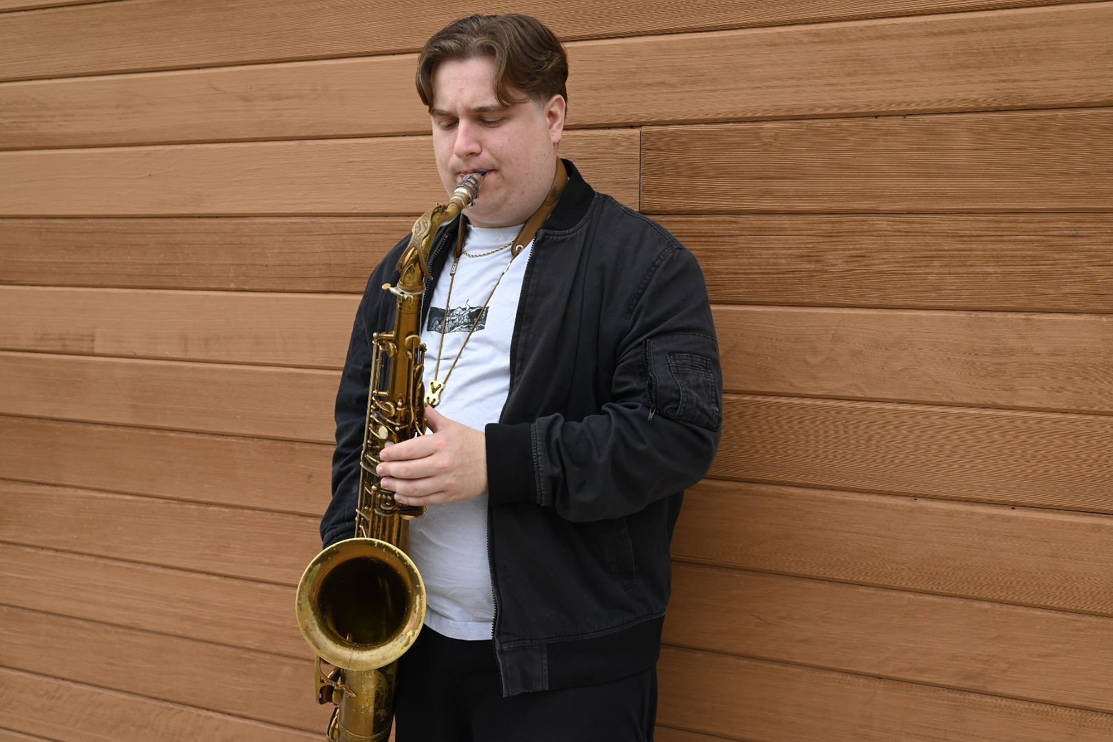 A young man with brown hair is standing against a wooden wall, playing a gold-colored tenor saxophone.