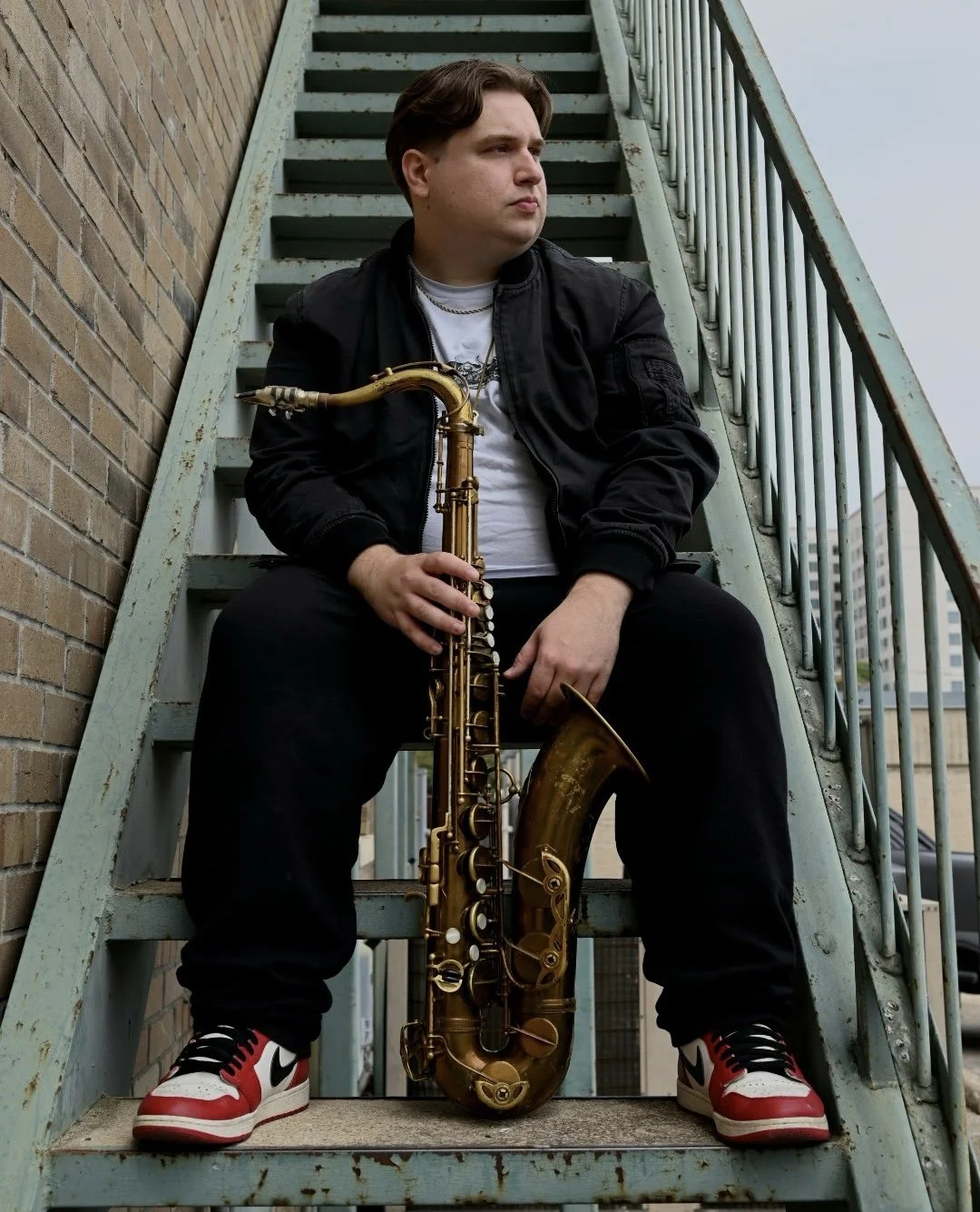 Young man sitting on outdoor metal staircase with a tenor saxophone between his legs, wearing a black jacket, white shirt, black pants, and red and white sneakers, looking to the side.