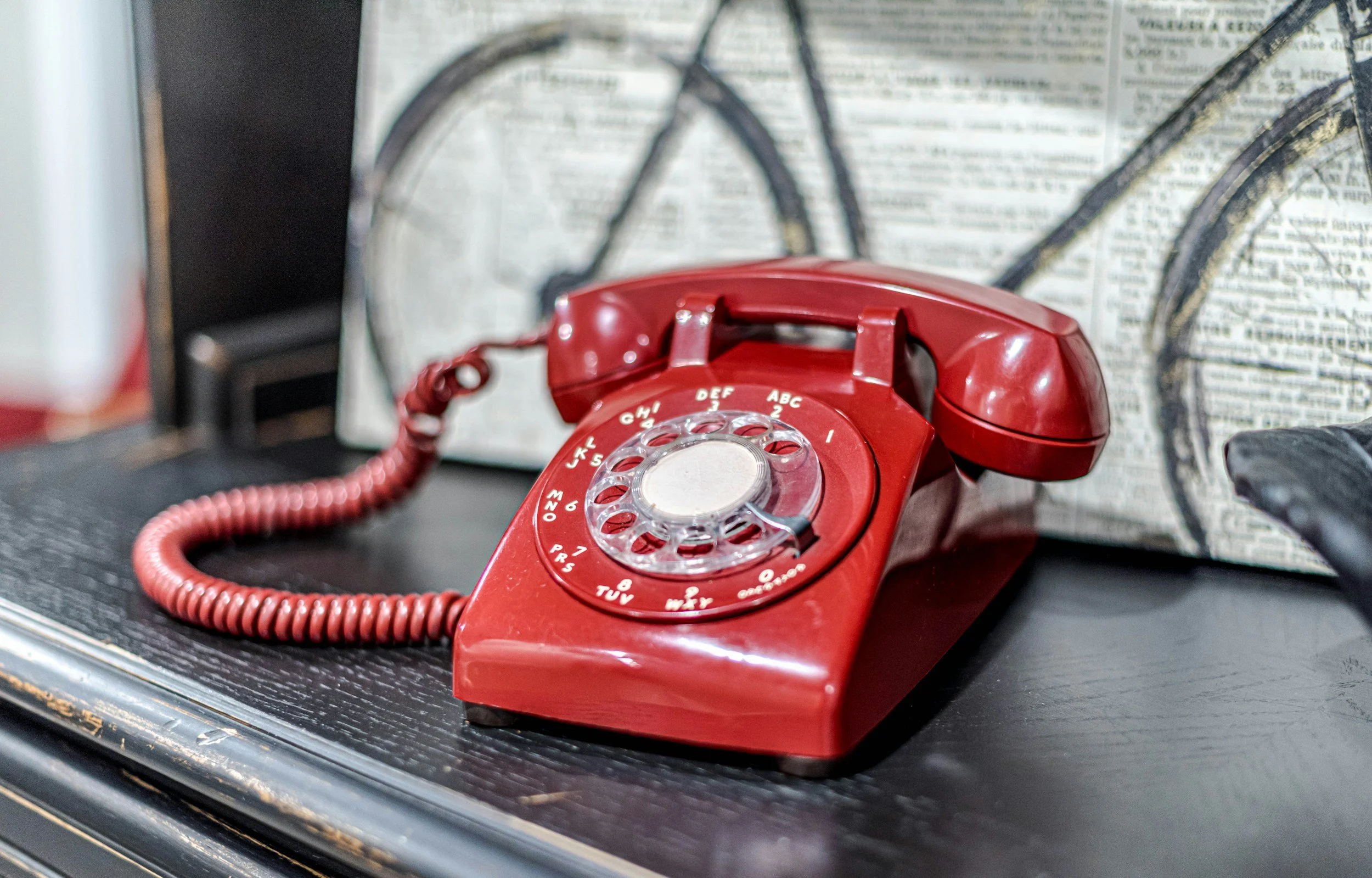 A red rotary dial telephone placed on a black surface with a blurred background including newspapers or printed material.