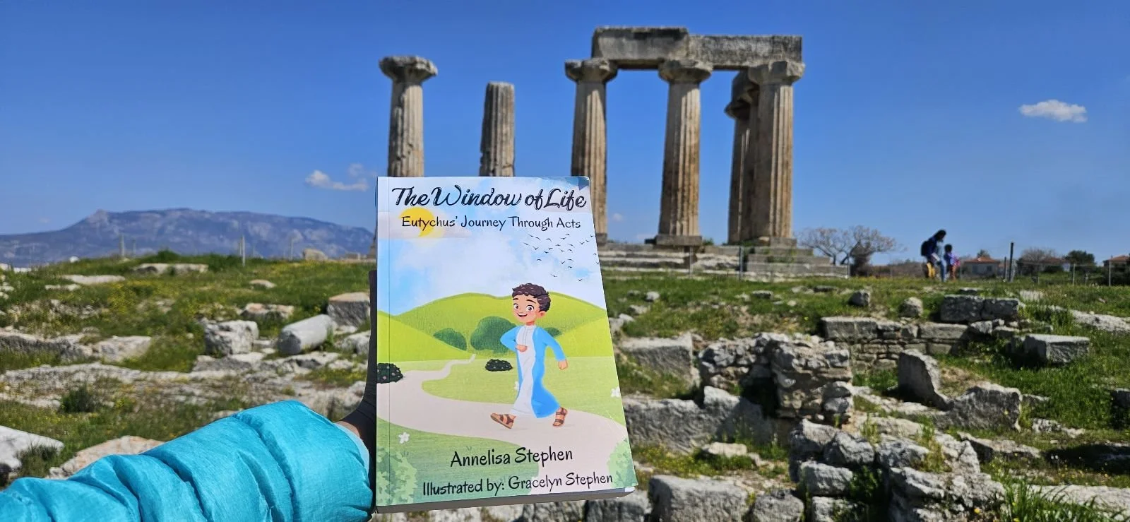 A person holding a children's book titled "The Window of Life" in front of ancient Greek ruins with three standing columns on a sunny day.