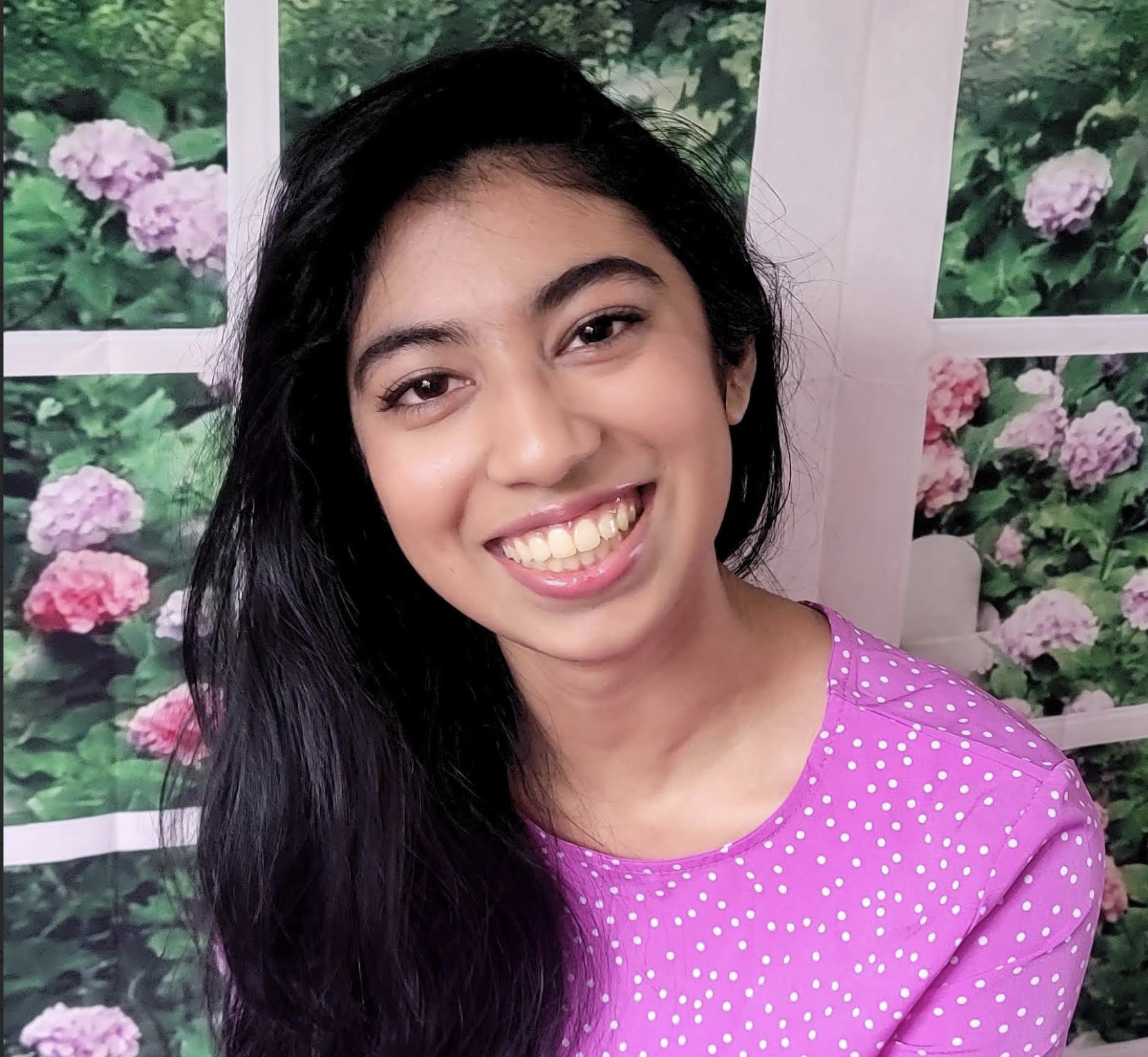 Smiling woman with long black hair wearing a pink polka dot dress, standing against a background of a wall with pink and purple flowers.