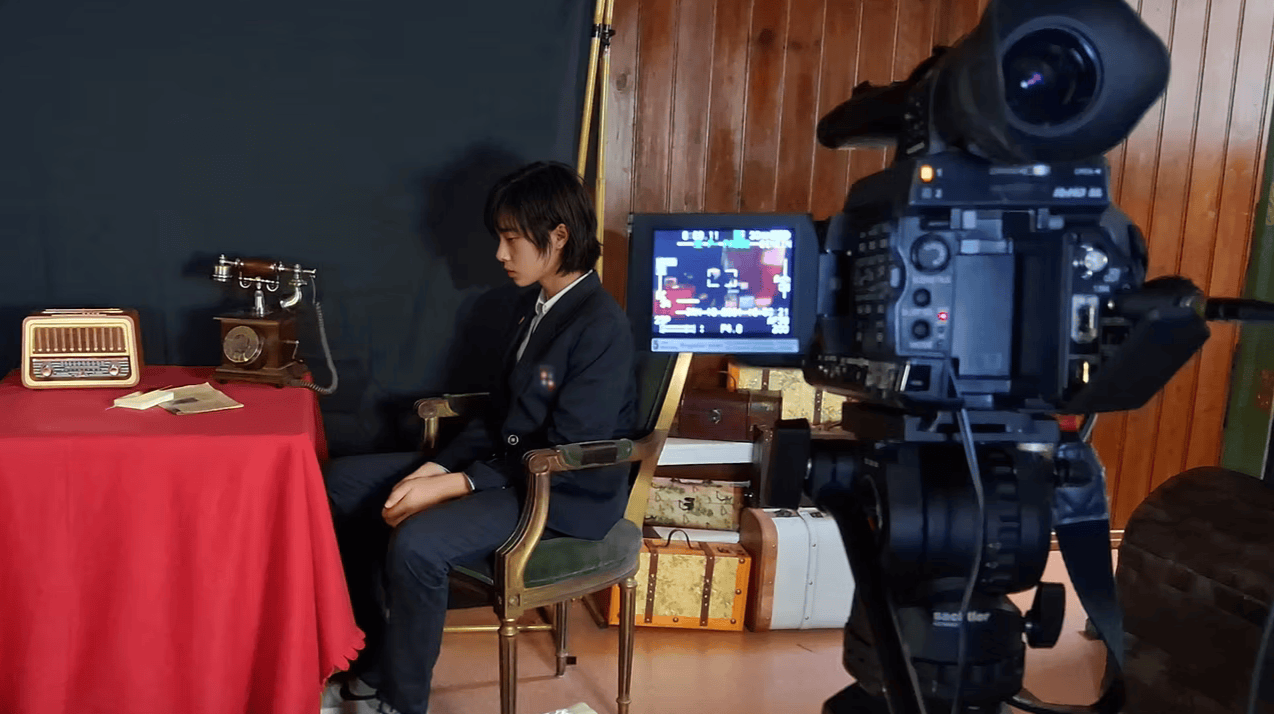 A young woman in a black suit sitting on a green and gold chair in front of a camera on a tripod. She is in a room with wooden walls, a table covered with a red cloth, an old-fashioned telephone, and a vintage radio. There are several packed suitcases on the floor behind her.
