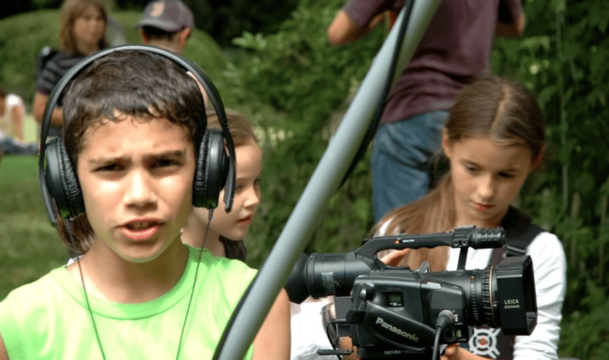 Children operating and observing a professional video camera outdoors, with a boy in a neon green shirt and headphones in the foreground.