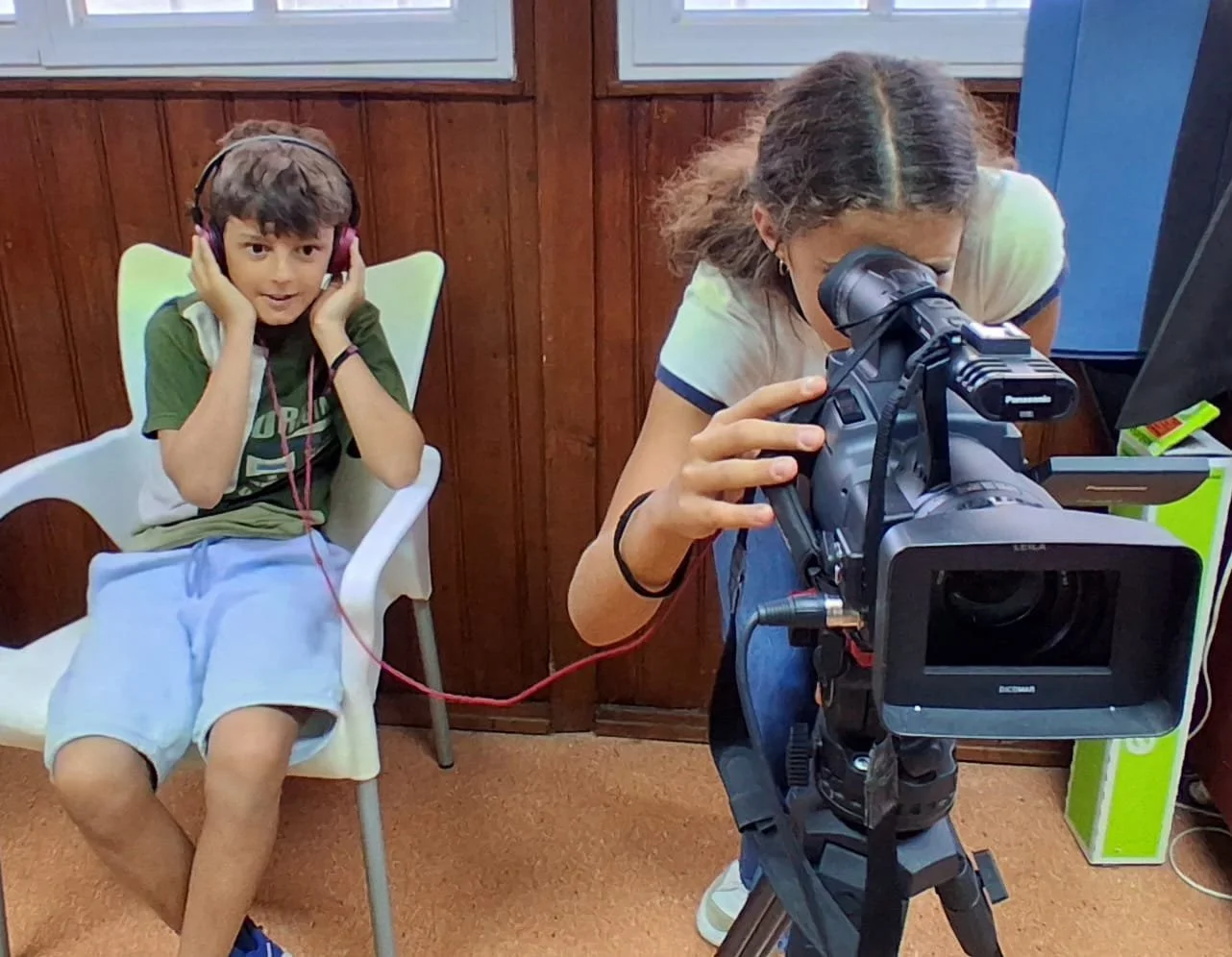 A young boy sitting on a chair with headphones on, while a woman looking through a camera or filming. The boy is smiling and appears to be listening to some audio, possibly for a recording or filming project.