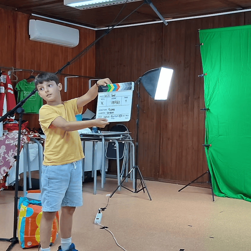 Young boy holding a film slate in a room with a green screen, studio lights, and a wooden wall.