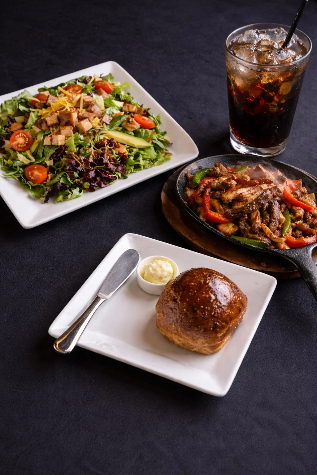 A meal with a side salad, a bread roll with butter, a sizzling stir-fry vegetable dish, and a glass of soda with ice on a black table.