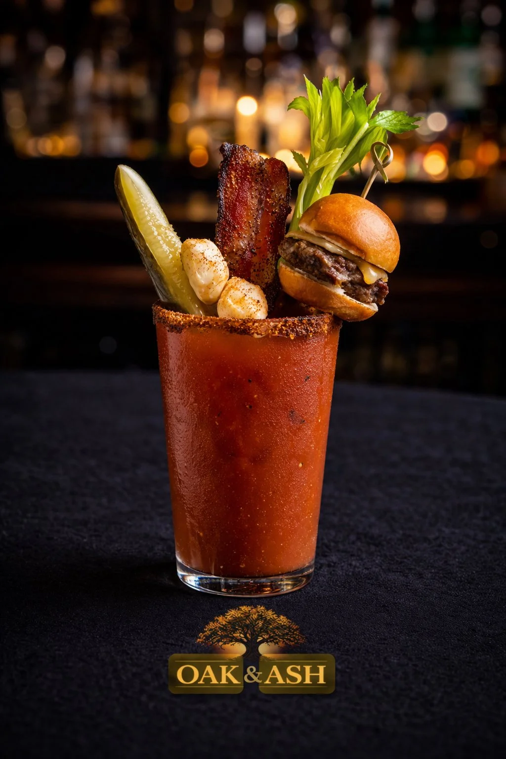 A cocktail glass filled with a red drink, garnished with a pickle spear, a strip of crispy bacon, a celery stalk, a small cheeseburger with a toothpick, and a couple of pearl onions, with a blurred bar background and Oak & Ash logo at the bottom.
