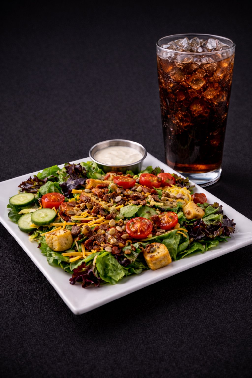 A plate of salad with cherry tomatoes, cucumbers, shredded cheese, lettuce, and grilled chicken, served with a side of dressing, alongside a glass of cola with ice.