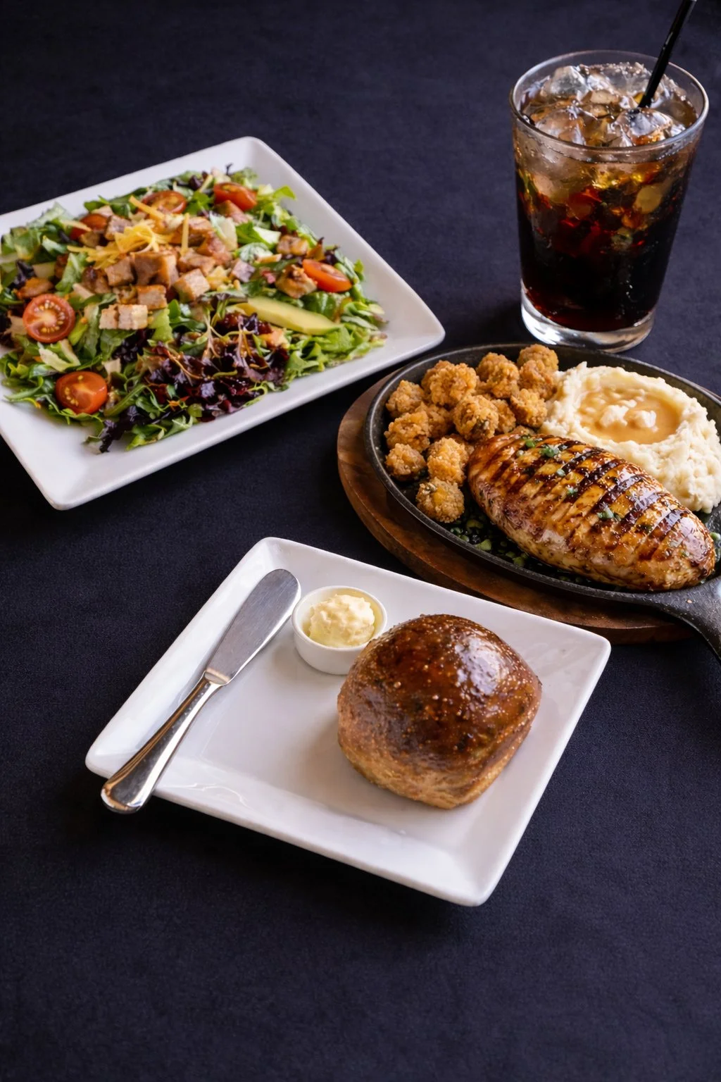 A meal featuring a side salad with greens, cherry tomatoes, cucumbers, and shredded cheese, a plate with bread roll and butter, a steak topped with bitter, mashed potatoes, fried chicken bites, and a drizzle of sauce, and a glass of cola with ice and