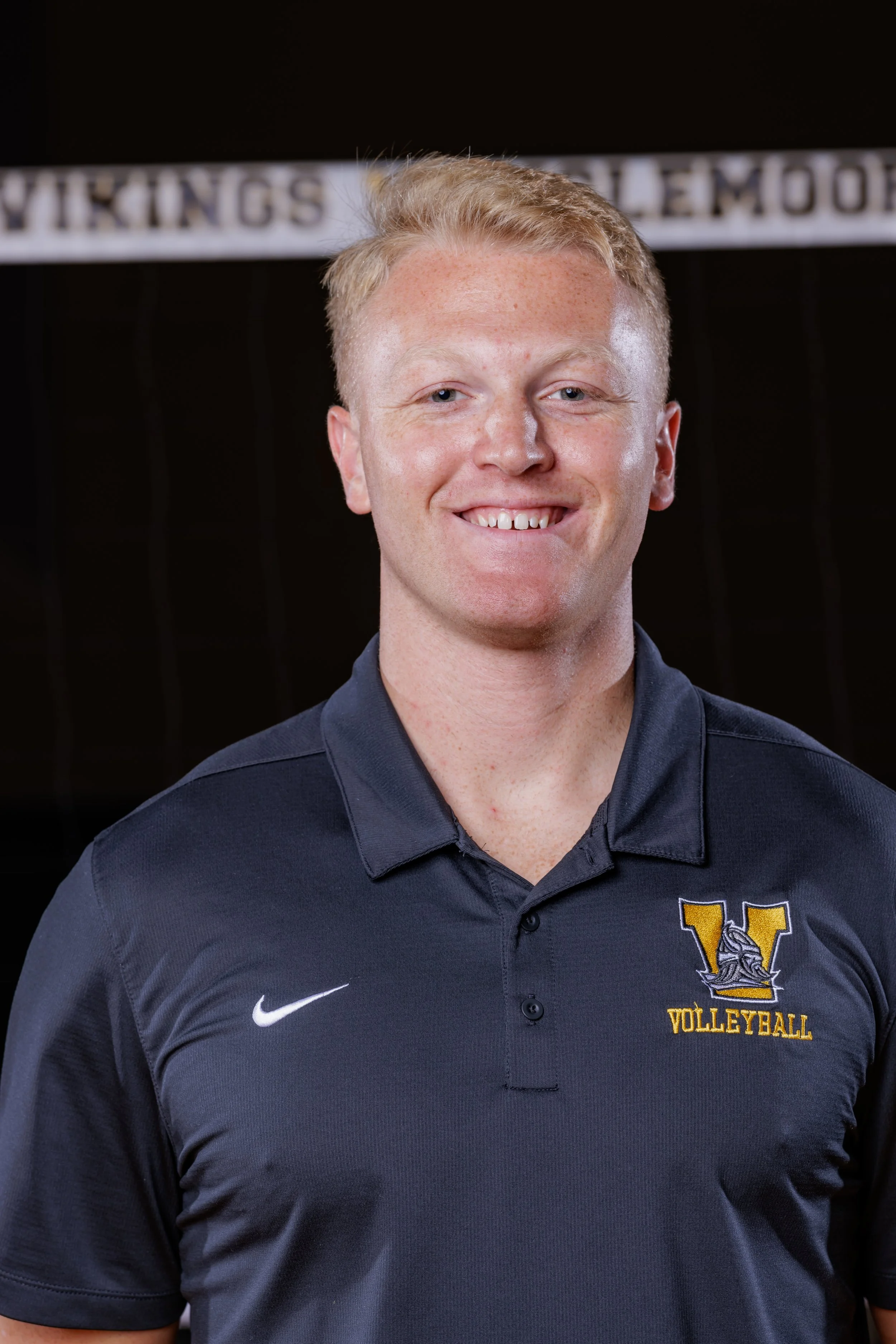 A young man with short blond hair smiling, wearing a dark navy volleyball team polo shirt with a yellow and black 'V' logo and the word 'VOLLEYBALL' embroidered on it, standing in front of a volleyball net with a sign in the background.