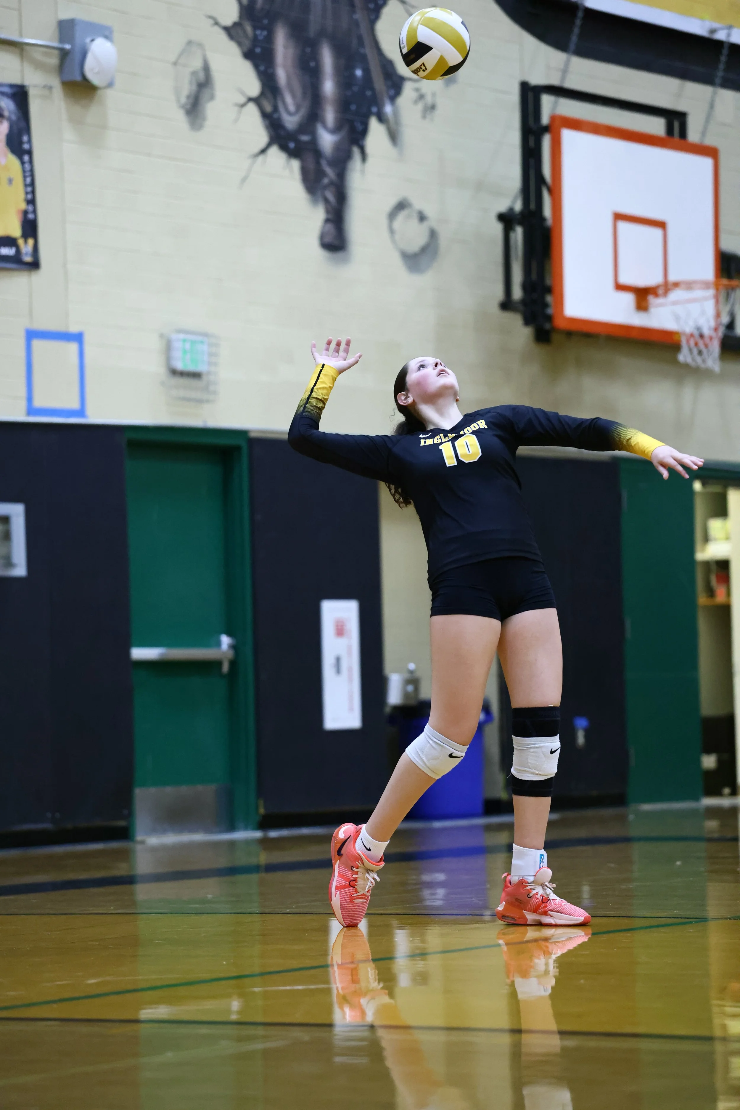 A female volleyball player with long dark hair tied in a bun, wearing a black and yellow uniform with the number 10, is inside a gymnasium. She is about to serve the volleyball, which is in midair above her head. The gym has a mounted basketball hoop, wall-mounted decor including a fish and sports posters, and reflective flooring.