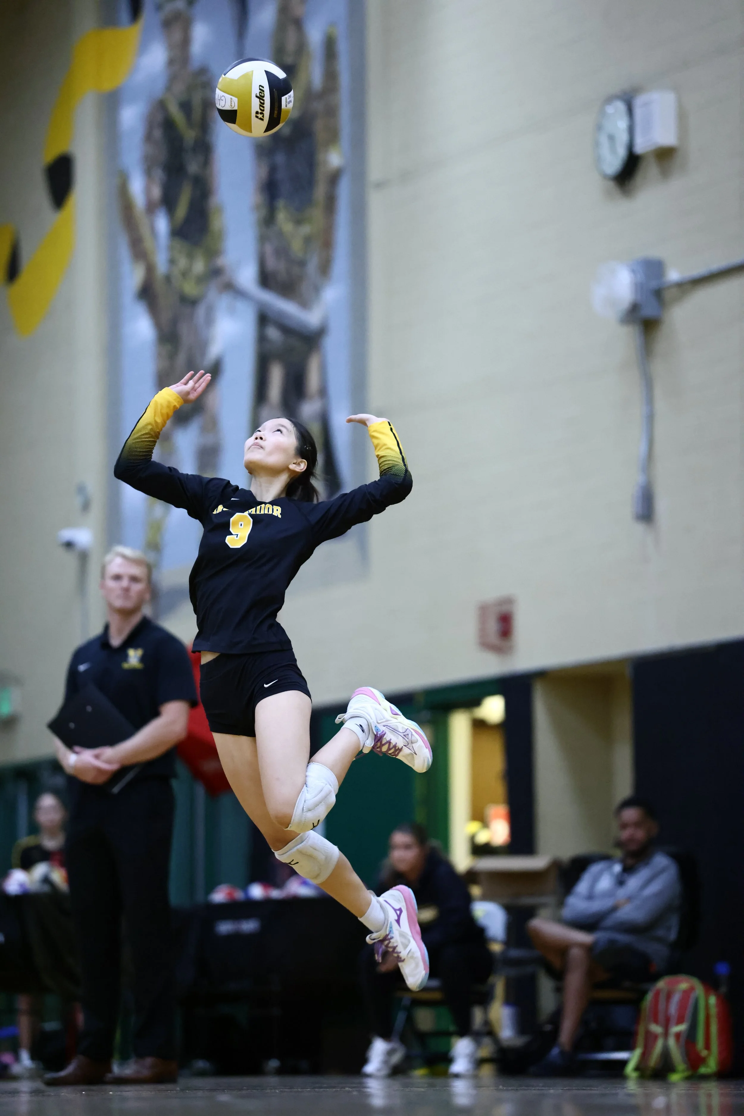 A female volleyball player in black and yellow uniform jumping to hit the ball during a match in an indoor gymnasium. Spectators and officials are visible in the background.