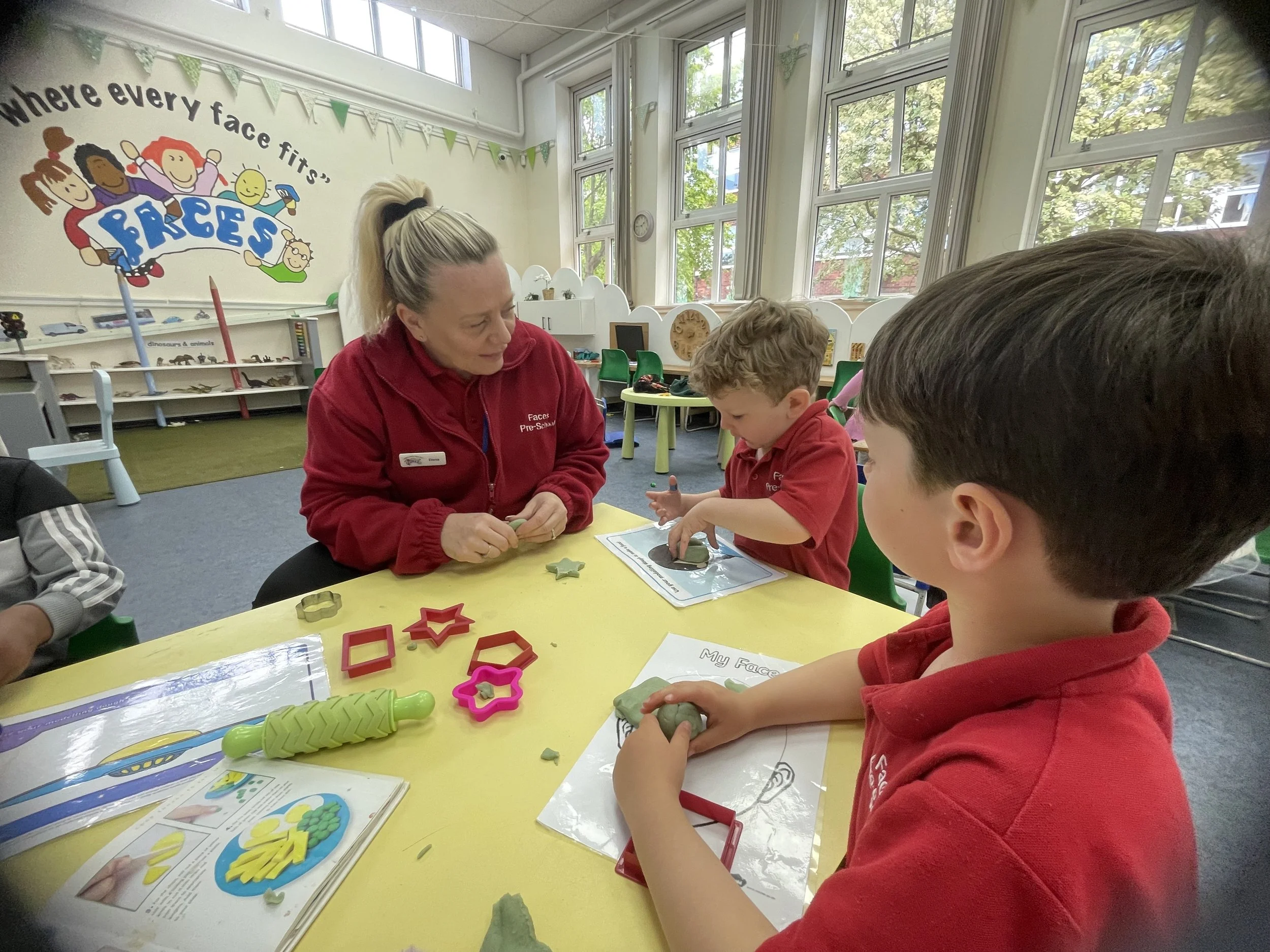 A teacher and children working on a crafting activity at a yellow table in a classroom with large windows. The classroom has a colorful sign on the wall that says 'Where every face fits' with illustrations of children, and various craft supplies on the table and shelves.