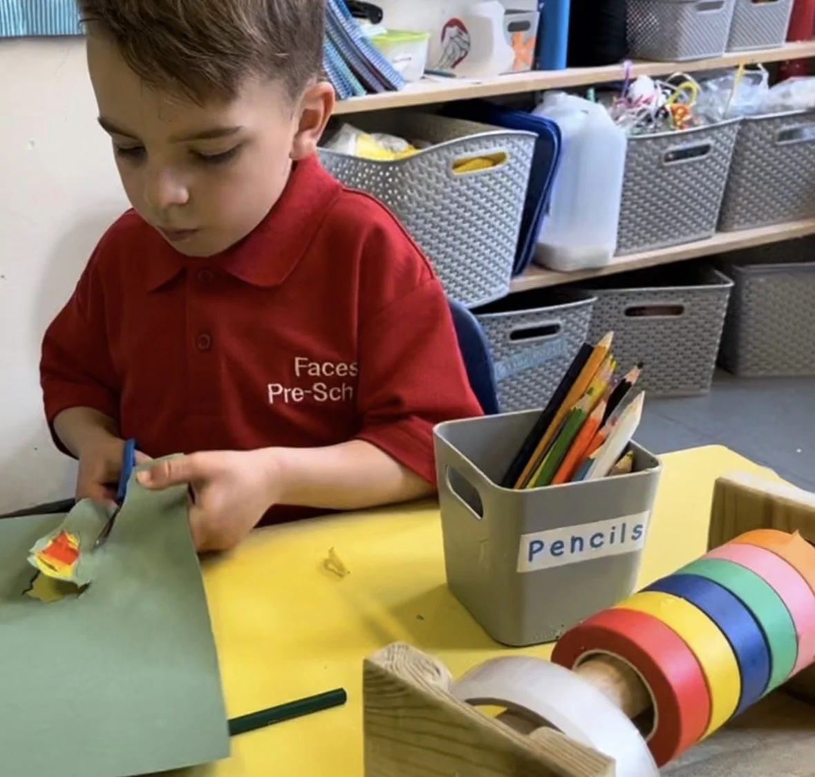 A young boy in a red shirt with 'Faces Pre-School' on it is cutting paper with scissors at a table in a classroom. On the table, there is a container labeled 'Pencils' filled with colored pencils, and a roll of colorful paper or tape.