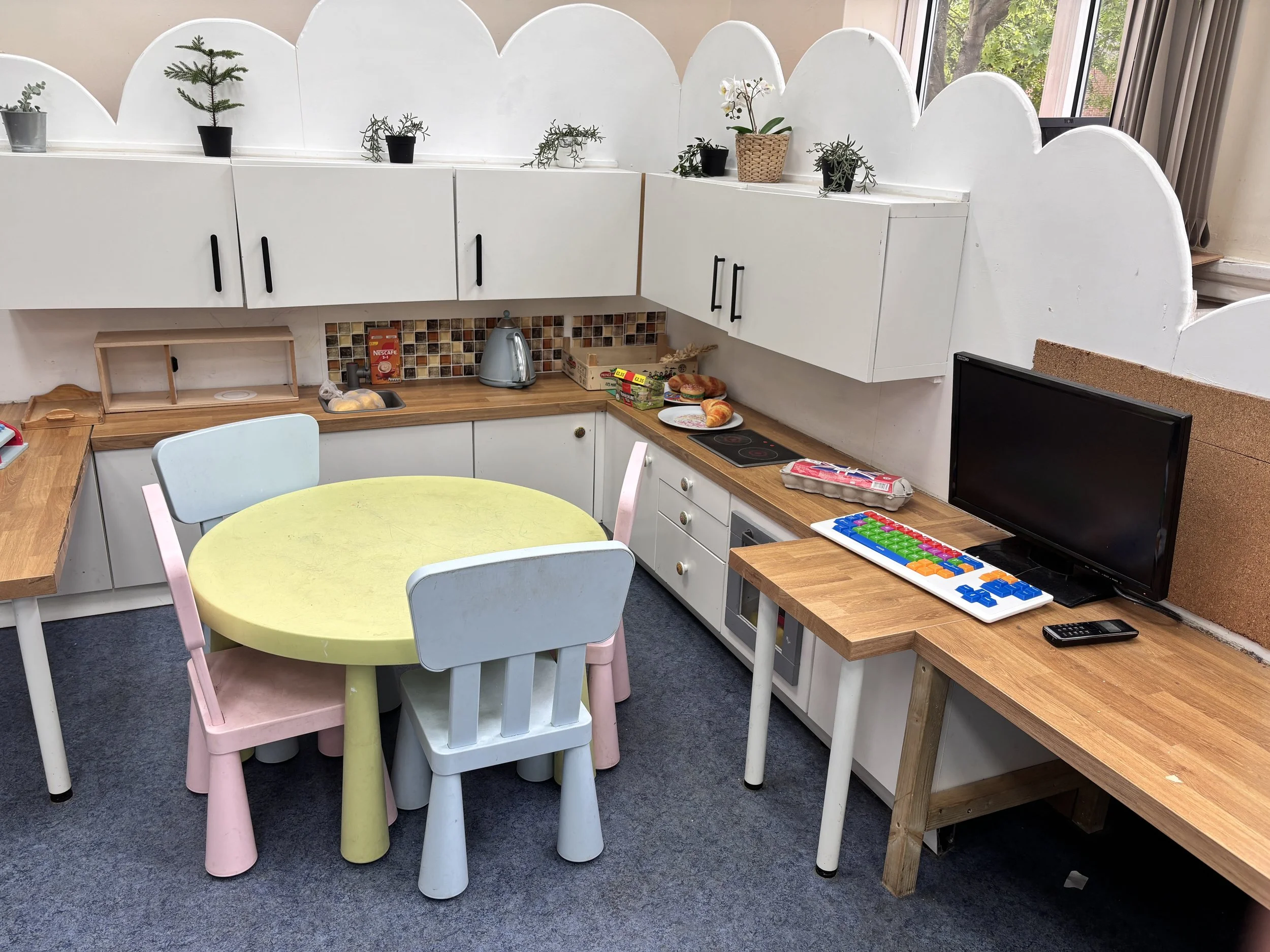 A kitchen with white cabinets, a round pastel yellow table with white and pink chairs, and various plants on top. A fan, some food items, and a TV are on the countertop. There is a window with curtains letting in natural light.