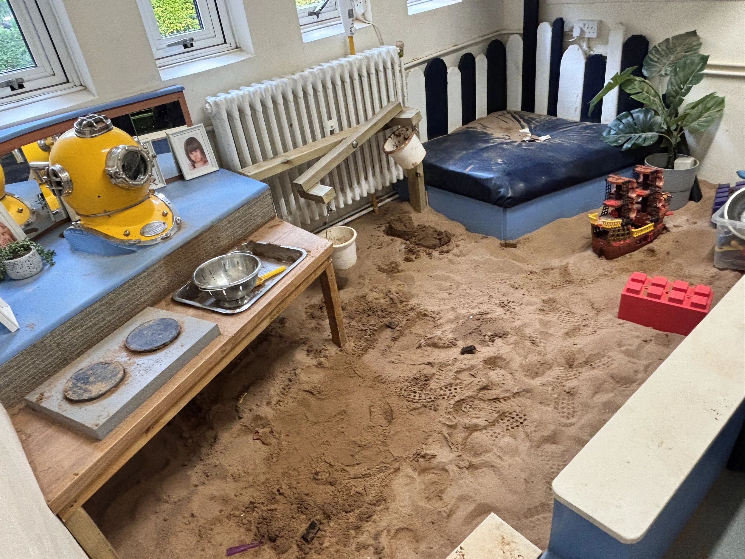 A room with sandy ground, a wooden bench, and a dining table with a yellow vintage diving helmet, framed photographs, a plant in a pot, a pile of toy bricks resembling a castle, and children's toys.