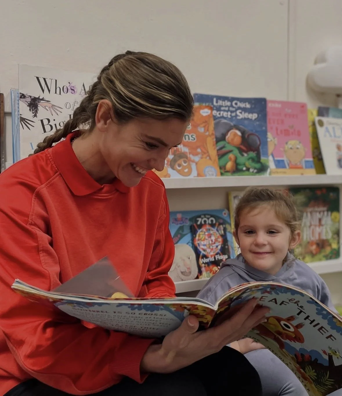 A woman in a Brentwood Preschool and a young girl reading a children's book together in a bookstore or library. The woman is smiling and wearing a red shirt, and the girl is smiling and wearing a gray hoodie.