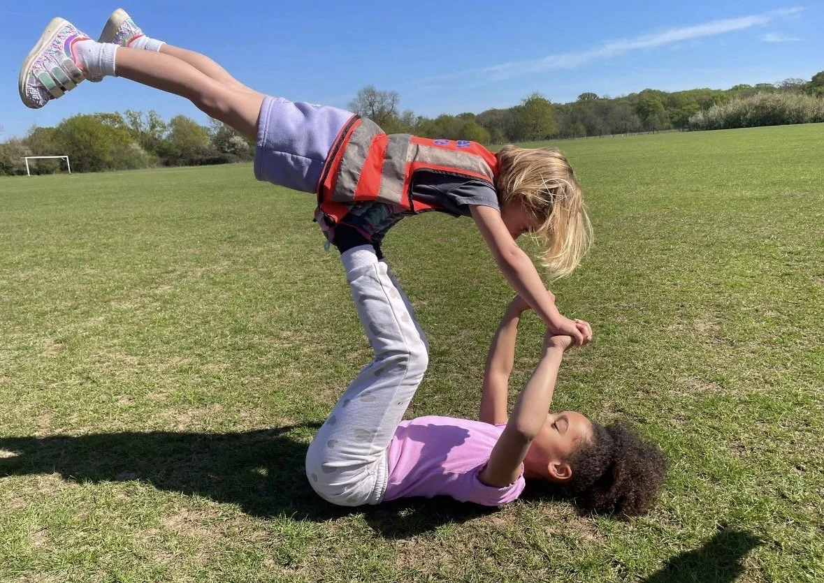Two young girls playing on a grassy field, in holiday club in Brentwood, one girl is lying on her back, and the other girl is balancing on her hands with her legs elevated, engaging in a playful handstand game.