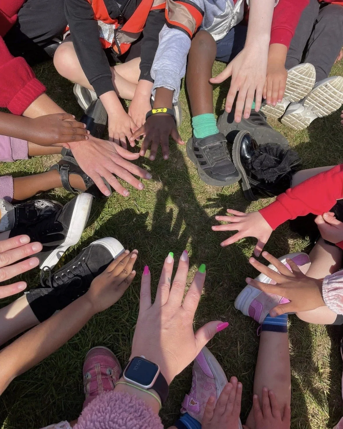 Children and adults sitting in a circle on grass, during their holiday club in Brentwood reaching towards the center with their hands together, displaying unity and teamwork.