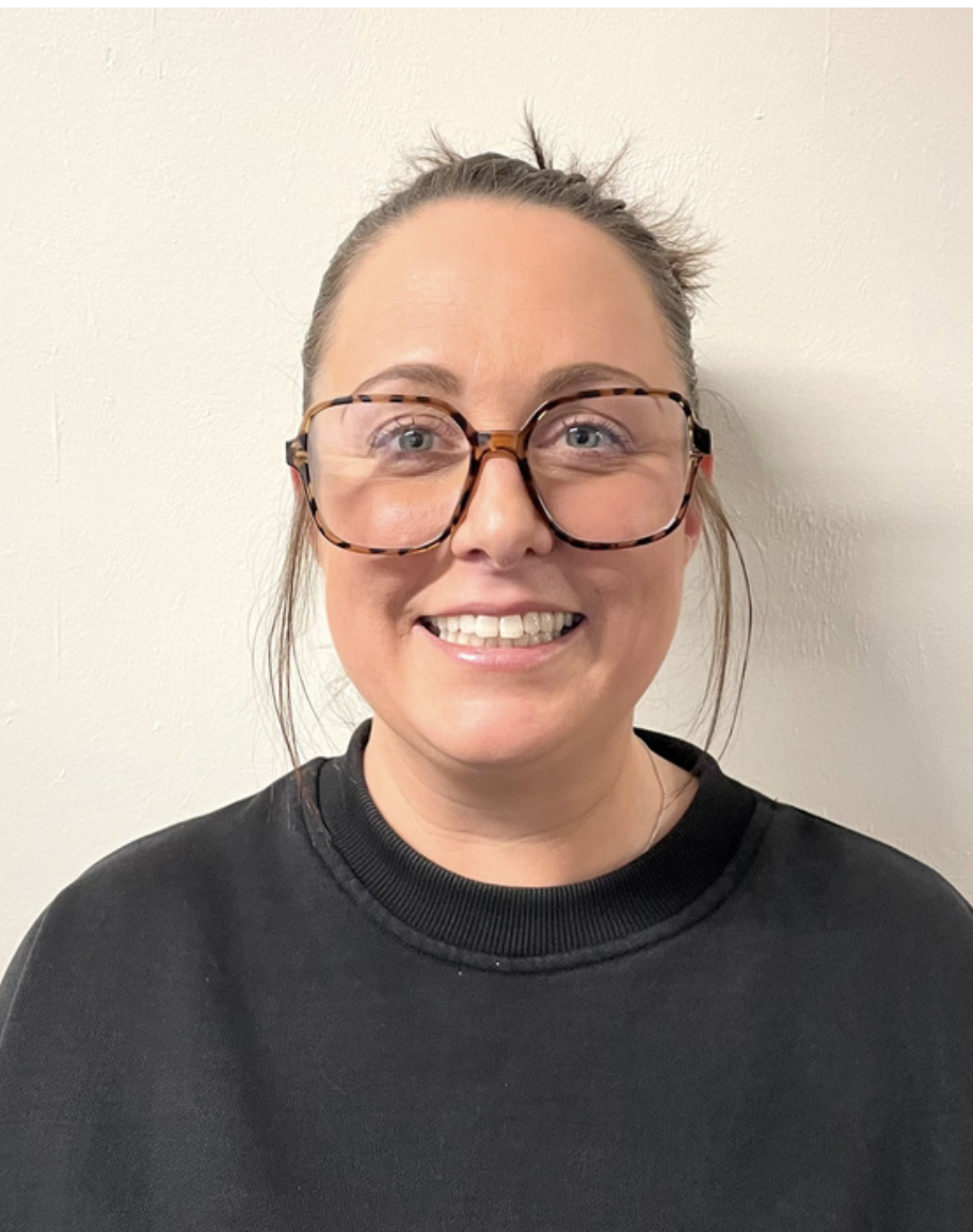 A Brentwood preschool playworker smiling with glasses and a black shirt standing in front of a plain white wall.