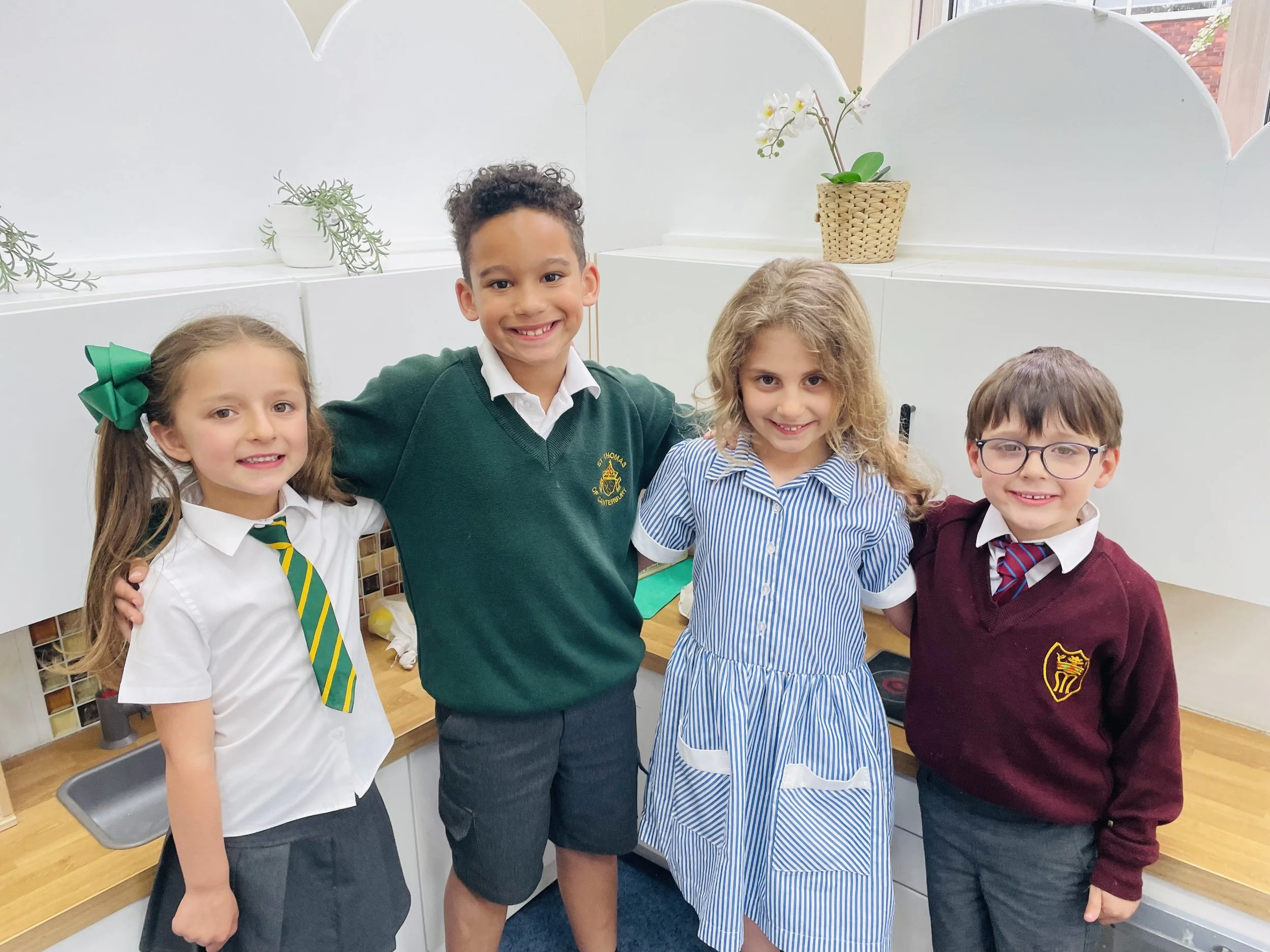 Group of four smiling children, two girls and two boys, standing indoors against a white backdrop with potted plants on a shelf.