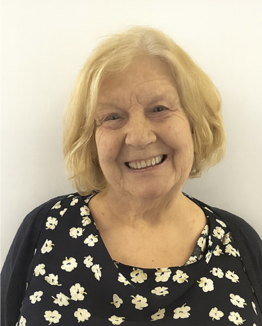 Smiling elderly woman with blonde hair wearing a black dress with white floral pattern, standing against a plain white background.