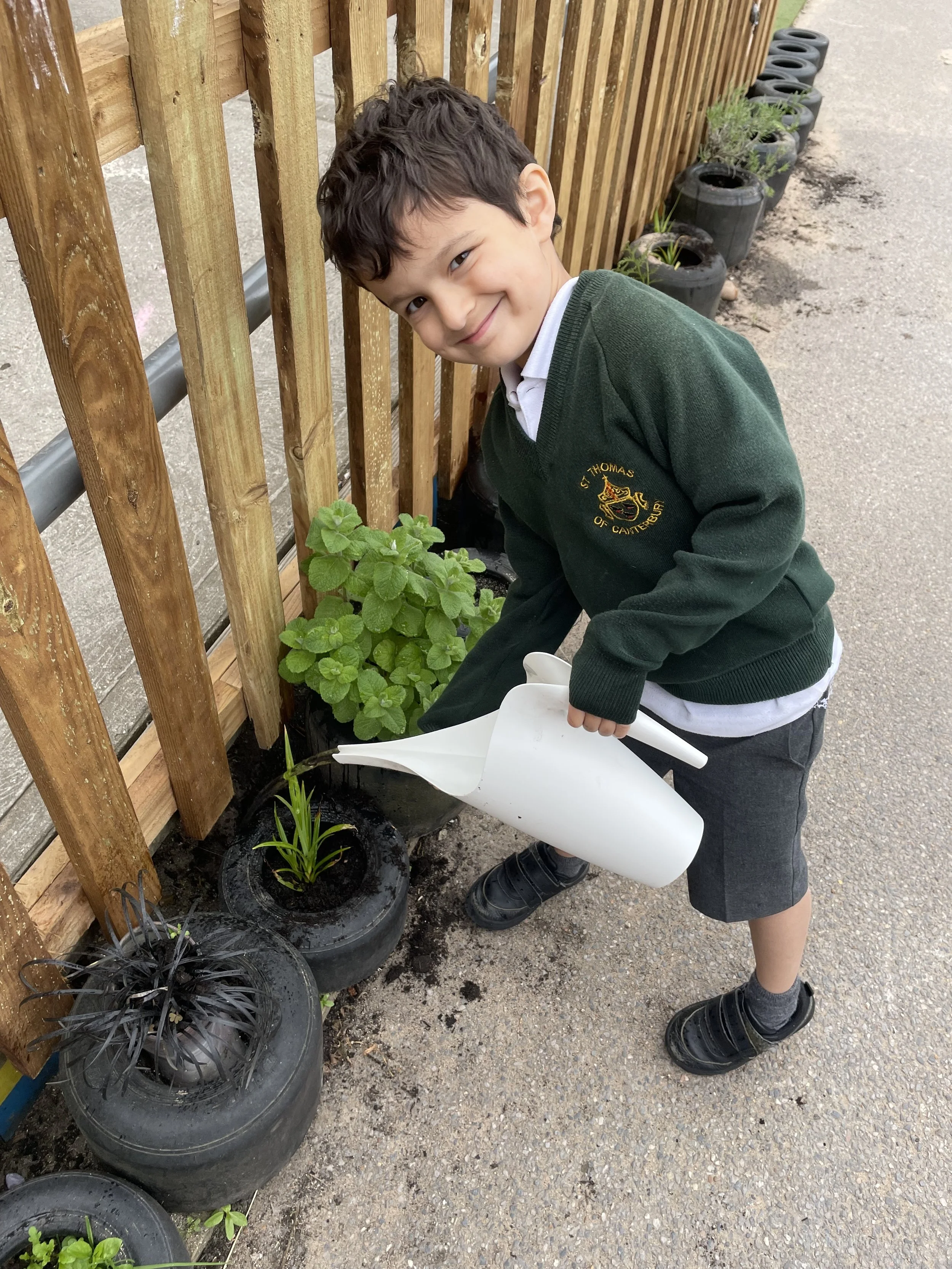 A young boy in a school uniform watering plants in black pots, standing on a paved surface next to a wooden fence with small plants growing in the background.