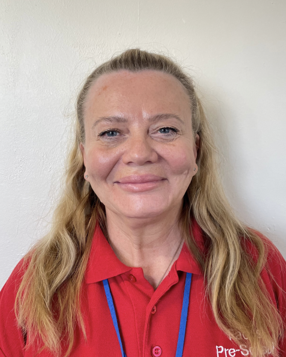 A preschool supervisor in Brentwood preschool with blonde hair, blue eyes, and a friendly smile wearing a red collared shirt with a lanyard, standing against a plain white wall.