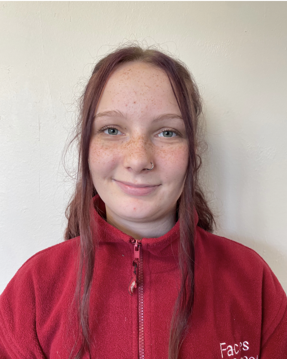 Young play worker from Brentwood Preschool with red hair, freckles, and a nose piercing, wearing a red zip-up fleece jacket, standing against a plain beige wall.