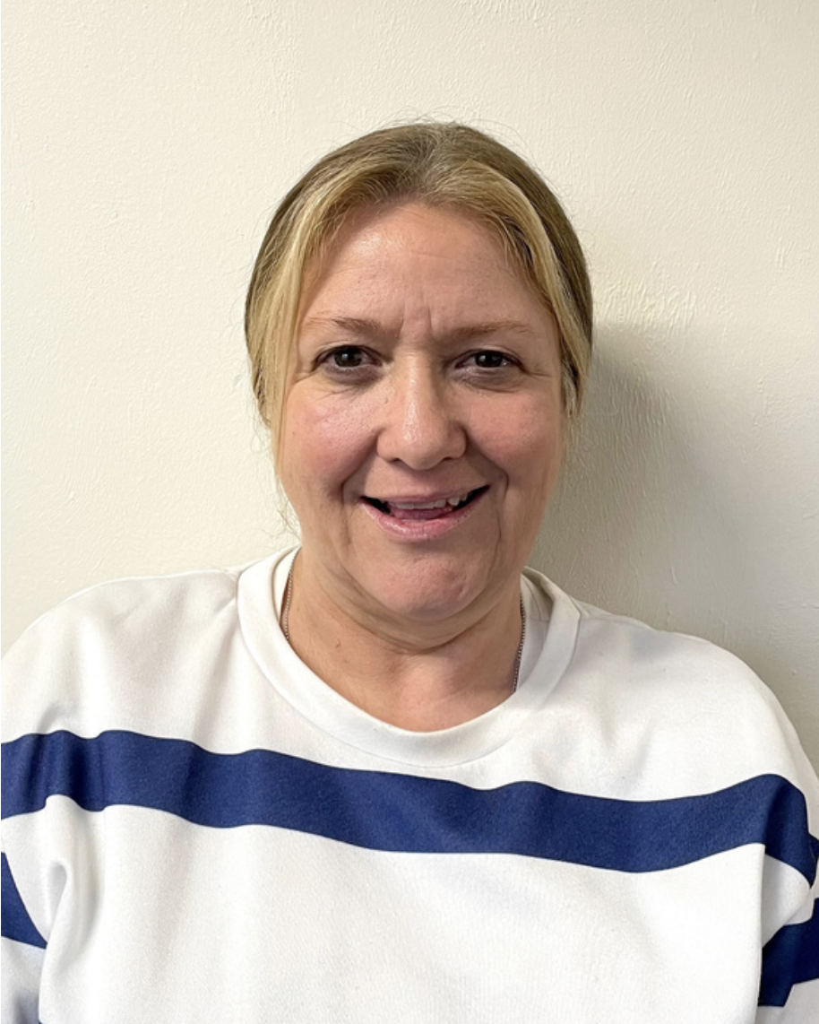 A Prechool playworker middle-aged woman with blonde hair wearing a white shirt with blue horizontal stripes, standing against an off-white wall.