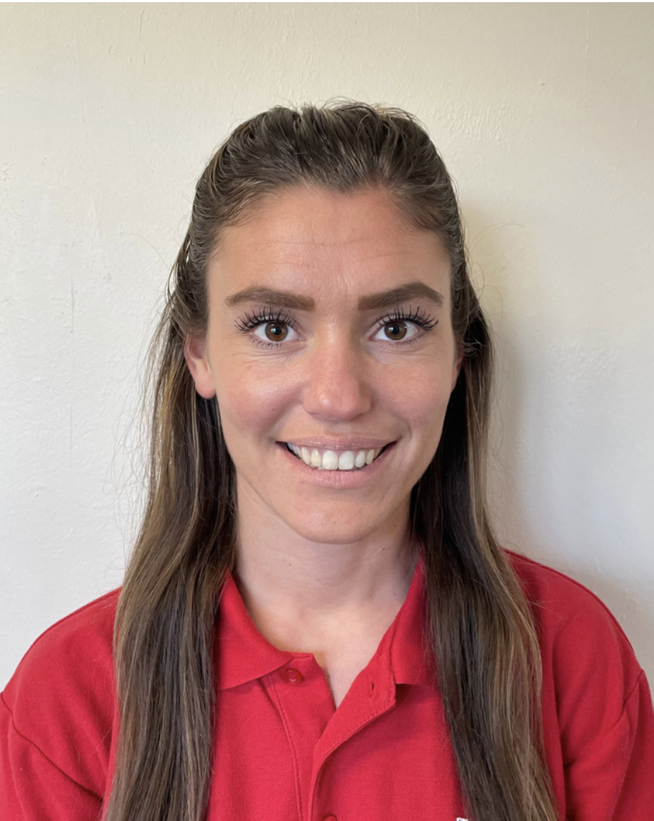 A play worker in Brentwood preschool with long brown hair, wearing a red shirt, smiling in front of a plain beige wall.