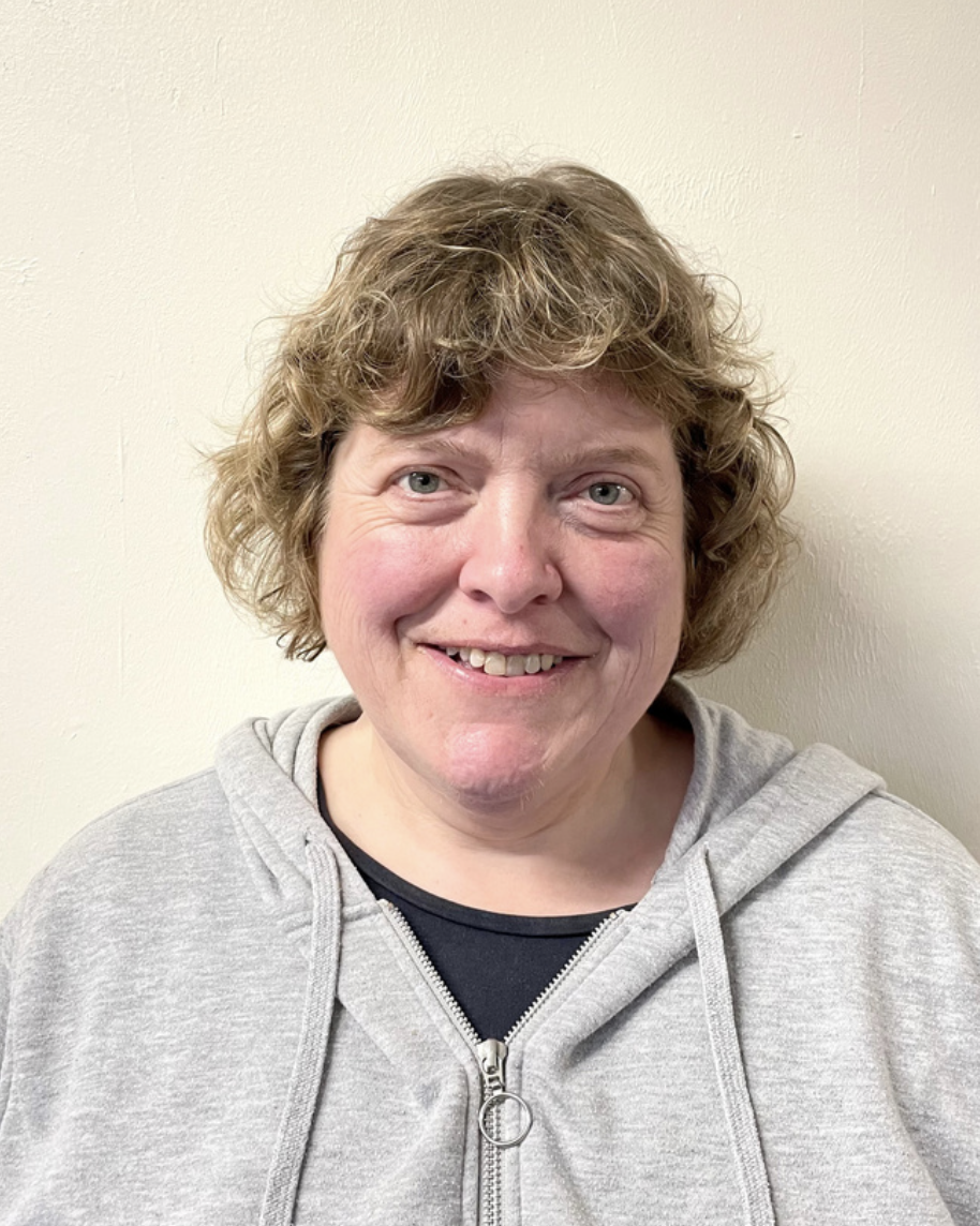 A woman with curly, light brown hair smiling at the camera, wearing a gray hoodie over a black shirt, standing against a plain white wall.