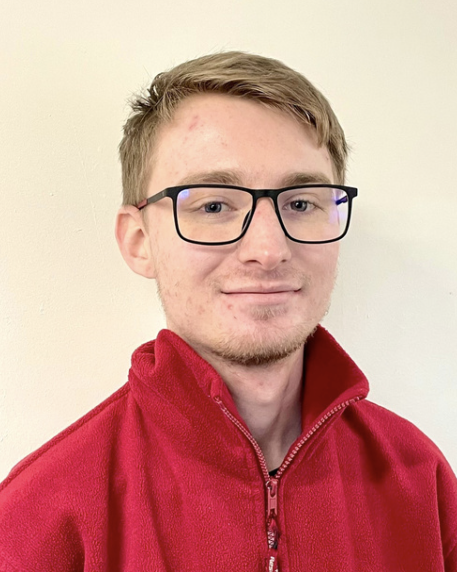 A young man with light brown hair, wearing glasses and a red zip-up fleece, standing against a plain light-colored wall.