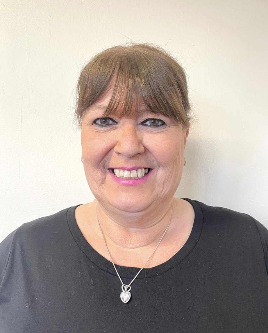 Smiling playworker from Brentwood preschool with short brown hair, wearing a black shirt and a silver heart-shaped pendant necklace, standing against a plain white wall.