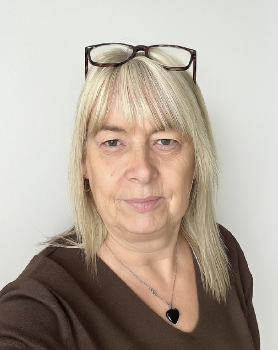 A supervisor from a Brentwood preschool with blonde hair and glasses resting on her head, wearing a brown shirt and a necklace with a black heart pendant, standing against a plain white background.