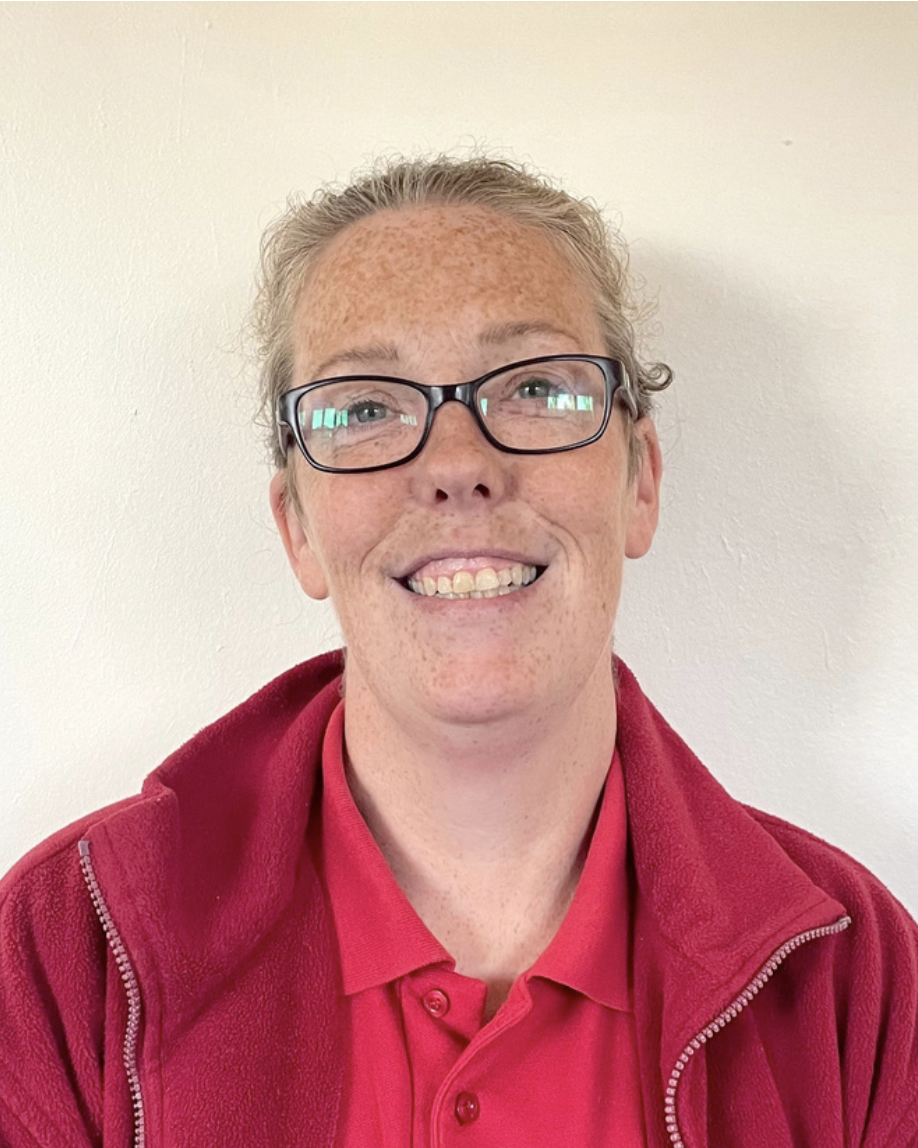 A play worker from Brentwood Preschool with glasses and freckles smiling at the camera, wearing a pink polo shirt and a red fleece jacket against a plain white wall.