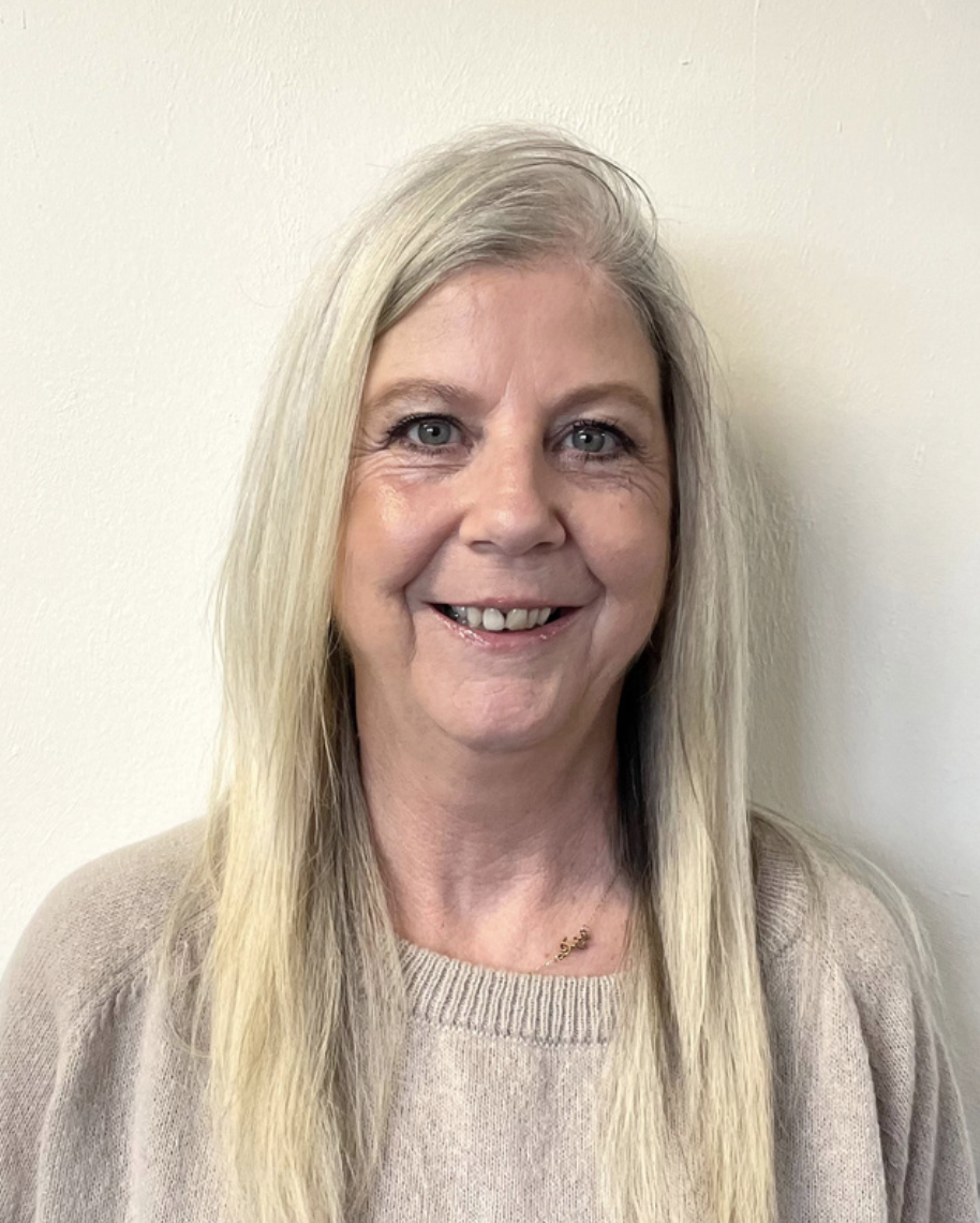 A woman with long gray hair, blue eyes, and a friendly smile, wearing a beige sweater and a small gold necklace, standing against a plain off-white wall.