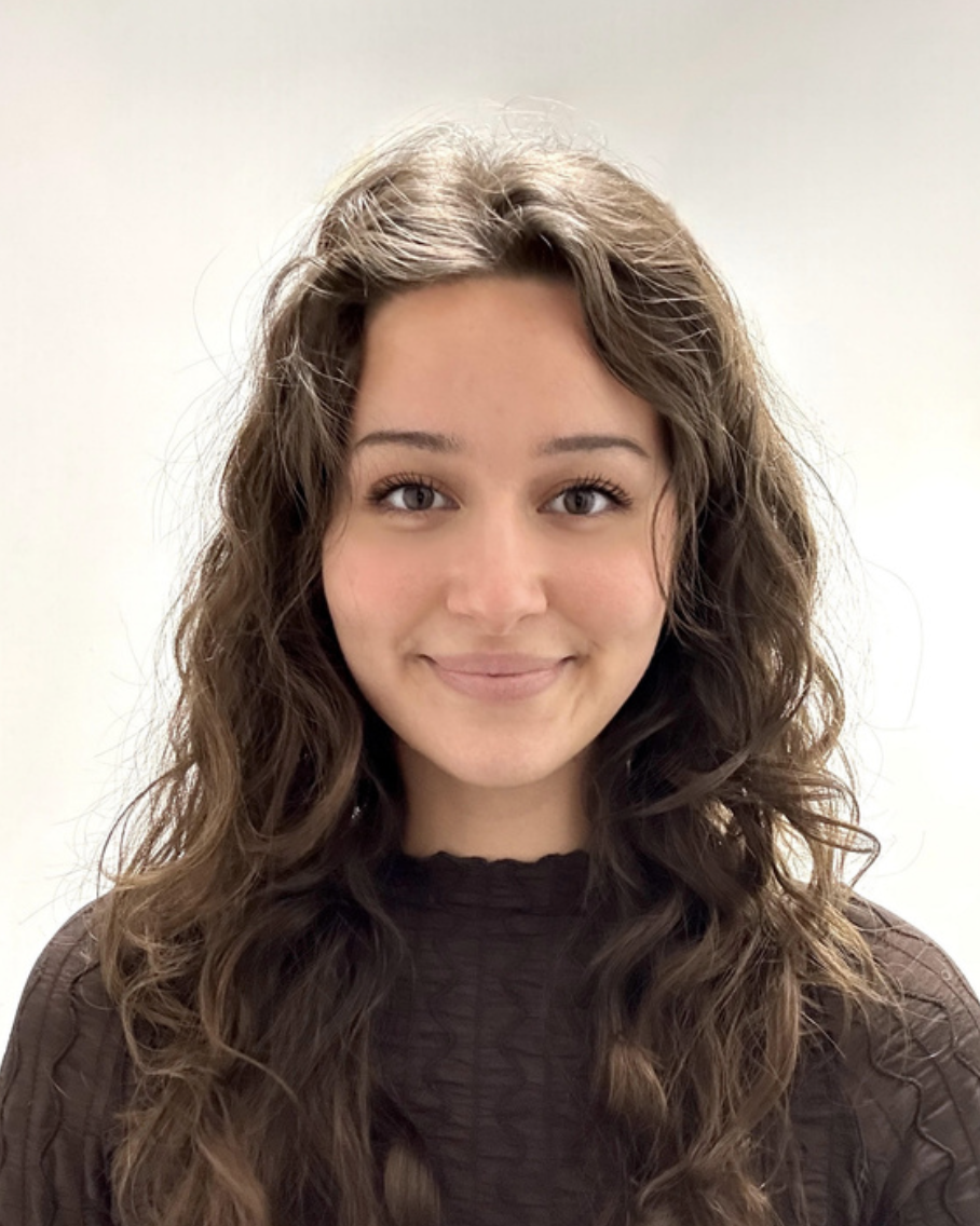 Portrait of a young woman with long, curly brown hair, from a Brentwood Preschool smiling, wearing a dark top, against a plain light background.
