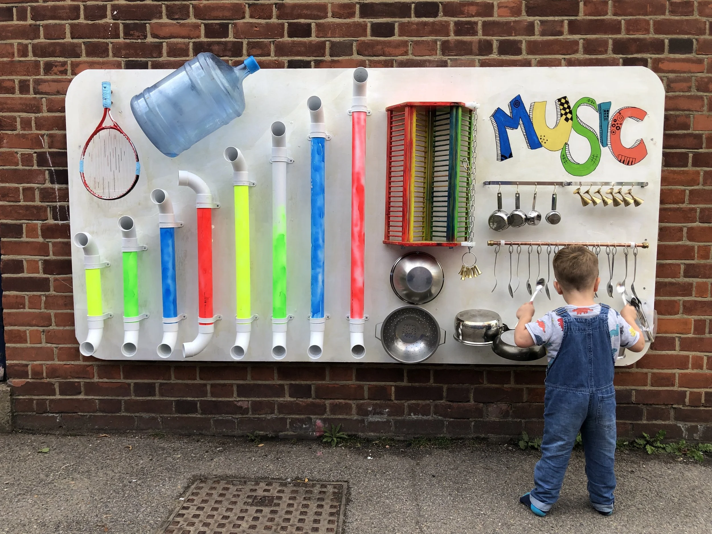 Child with short blonde hair, wearing denim overalls, standing outdoors a red badminton racket with blue handle tape, utensils, and kitchenware. The pipes are arranged vertically with glowing colors at the bottom, some connected to a large water jug. The child is facing the board with their back to the camera.